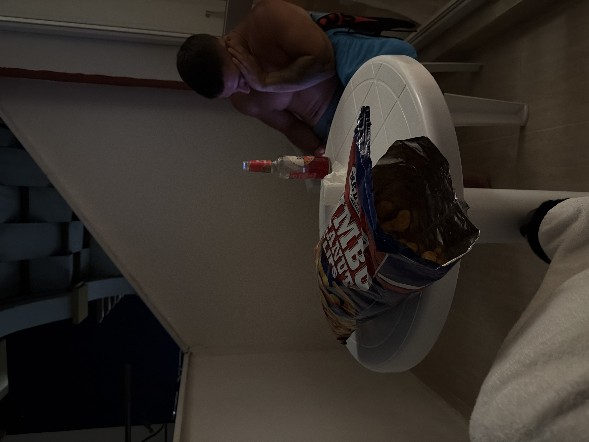 In Nesebar, Bulgaria, a man is seated at a round white table on a balcony. He is wearing blue shorts and is shirtless. His left arm is bent, and his hand is resting on his cheek, suggesting he is tired or bored. A bottle of beer and a large bag of chips are on the table. The chips are partially spilled out of the bag. The balcony appears to be part of a hotel or residential building, with the dark night sky visible in the background. The lighting is dim, creating a subdued atmosphere.