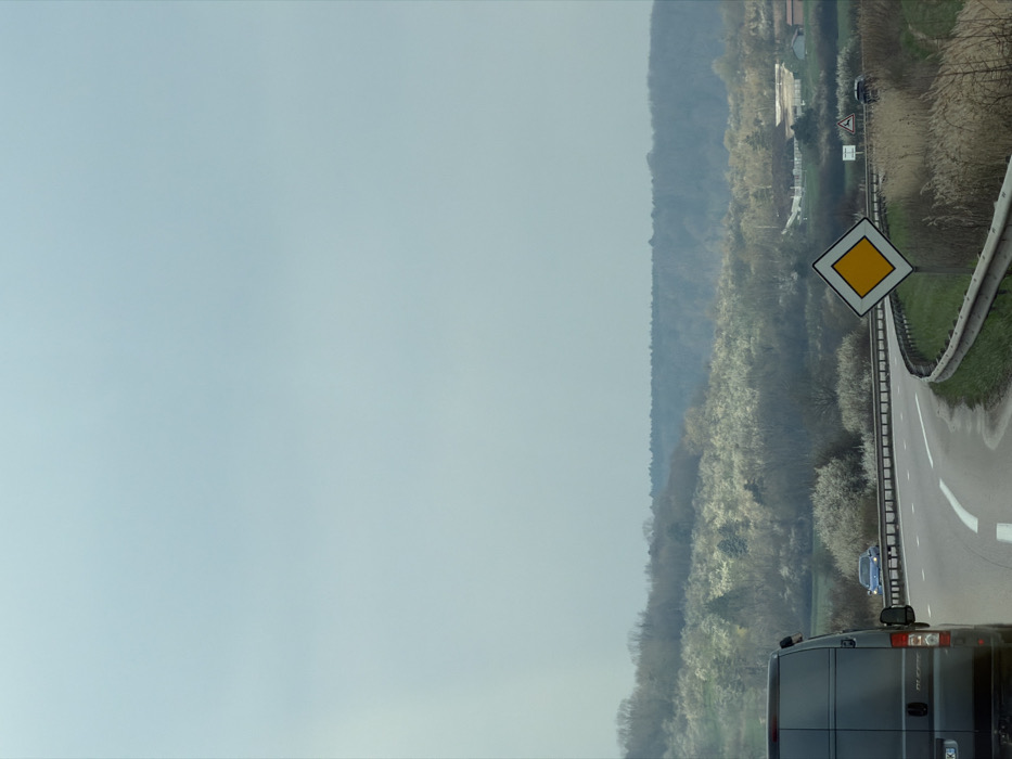 The image displays a rural road scene near Toul, France, under an overcast, light blue-gray sky. In the foreground, the rear of a dark panel van, possibly a Mercedes-Benz Sprinter variant, is visible on the lower left, displaying a partially visible European license plate ending in "KJ". To its right, an asphalt road curves sharply to the viewer's right, delineated by white solid and broken lane markings. A yellow diamond-shaped priority road sign with a white border is positioned on the right side of the road, beside a metallic guardrail that follows the curve, bordering an area of dry, tall grasses. Further along the curved road, a blue car with illuminated headlights is visible, heading away from the viewer. In the midground, a landscape of rolling hills is densely covered with a mix of deciduous trees, many displaying profuse white blossoms, characteristic of spring. Structures with light-colored roofs are discernible among the trees on the right side of the midground, with a darker band of forest forming the distant horizon. A dark SUV or car is partially visible near another, smaller road sign further in the distance on the right. No individuals are visible.