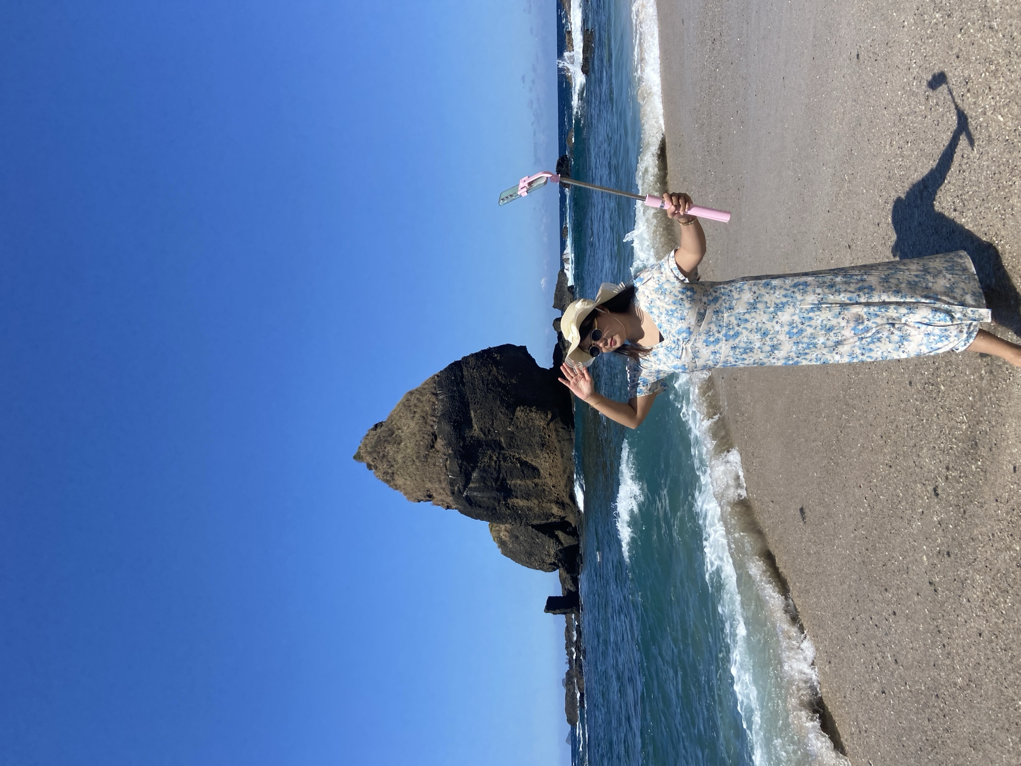 A person stands on a sandy beach, holding a pink selfie stick, and waving with their right hand. They are wearing a straw hat, sunglasses, and a floral-patterned dress. To the left of the person, there's a large, dark-colored rock formation rising from the ocean, while the ocean waves wash onto the beach. The sky is clear and blue. The shadow of the person and the selfie stick are visible on the sand. The location is Unknown, Indonesia.