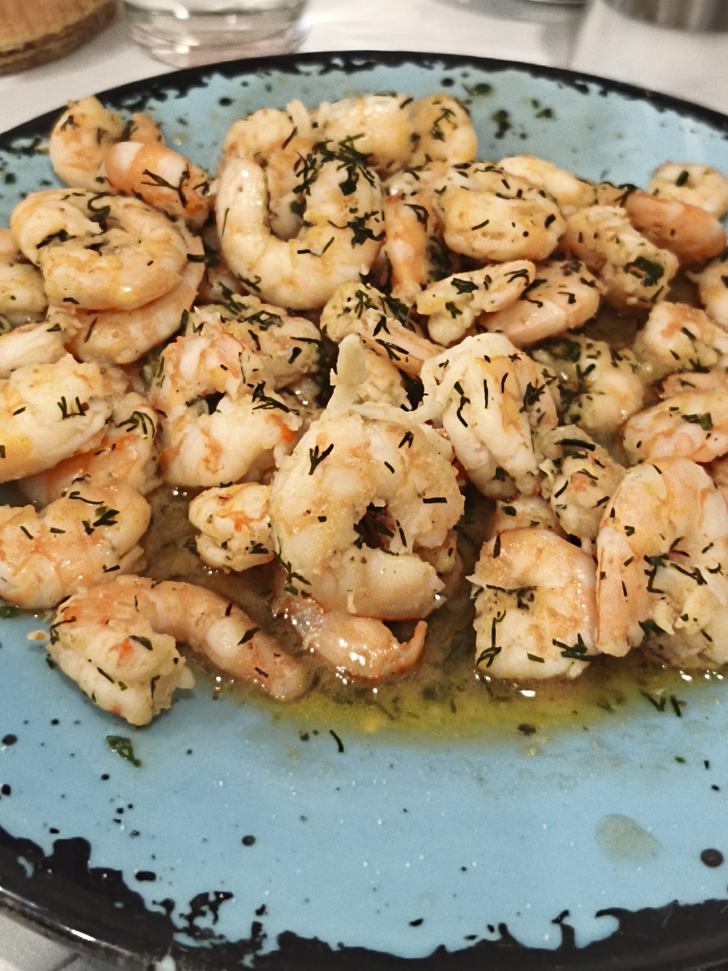 The image shows a blue plate filled with cooked shrimp, seasoned with what appears to be dill or another herb. The shrimp are bathed in a light-colored sauce, likely oil-based. The plate has a mottled blue surface with dark splatters. The location is not discernible from the image. There are no people visible. The scene depicts a close-up of the food item.