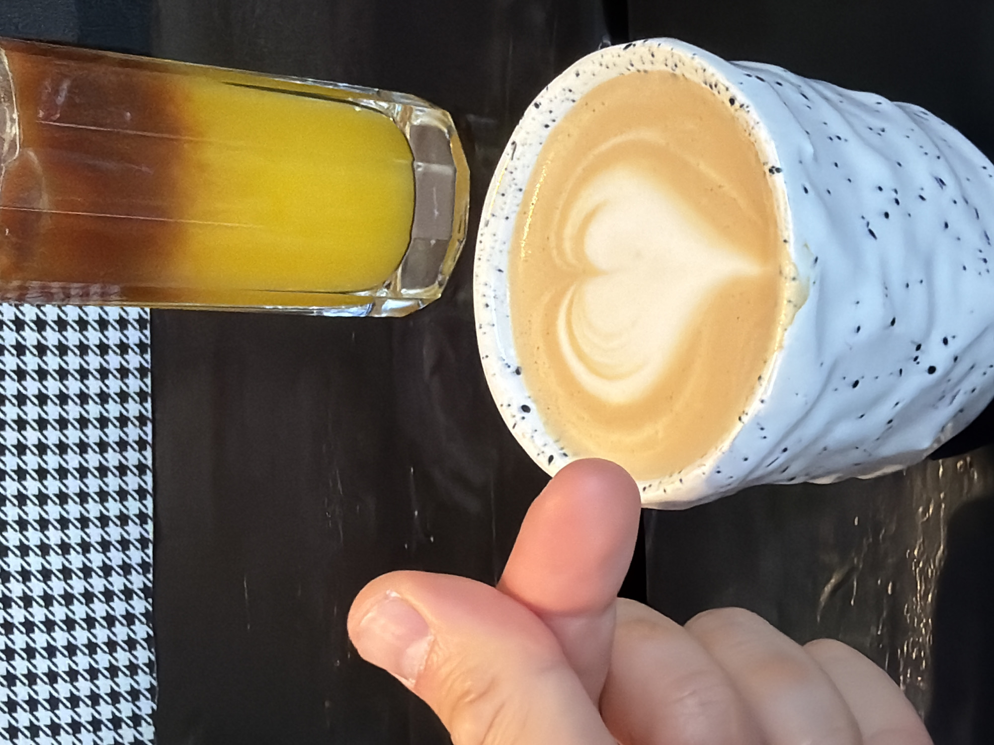 The scene depicts a glass of a yellow and brown layered beverage, possibly juice and coffee, alongside a white speckled mug containing a latte with a heart-shaped foam design. A hand is visible, making a finger gesture. The background consists of a black surface and a section with a black-and-white houndstooth pattern. The location in the city of Krasnoyarsk, Russia, cannot be determined from the visual data.