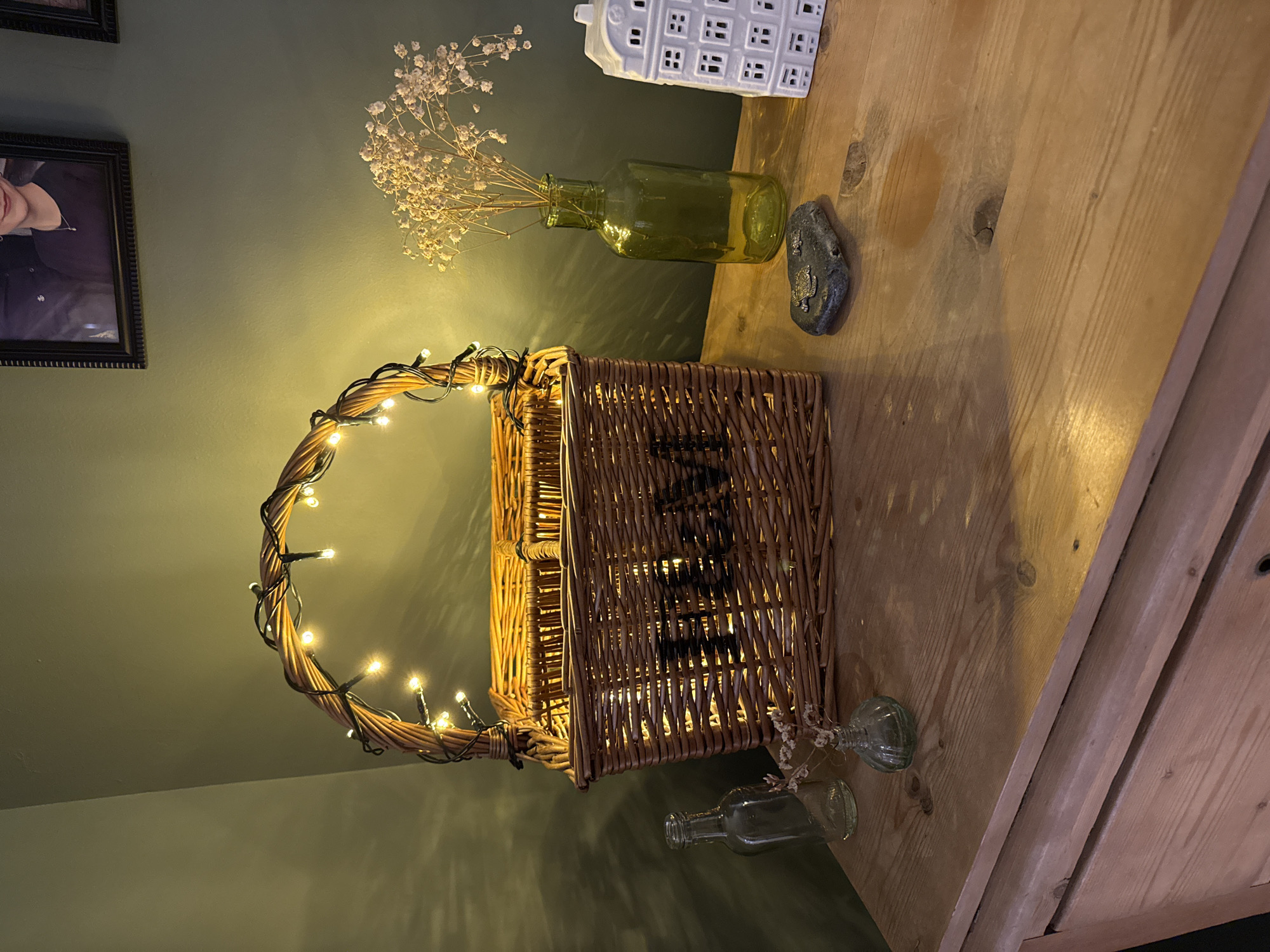 A wicker basket with fairy lights is placed on a wooden surface. Behind the basket are a small green glass bottle with dried flowers and a small white ceramic house-shaped ornament. The background is a dark green wall with a framed picture. Two small clear glass bottles are visible near the basket's base. The scene appears to be indoors, potentially in a room with a rustic or vintage decor style. The picture was taken in East Suffolk, United Kingdom.