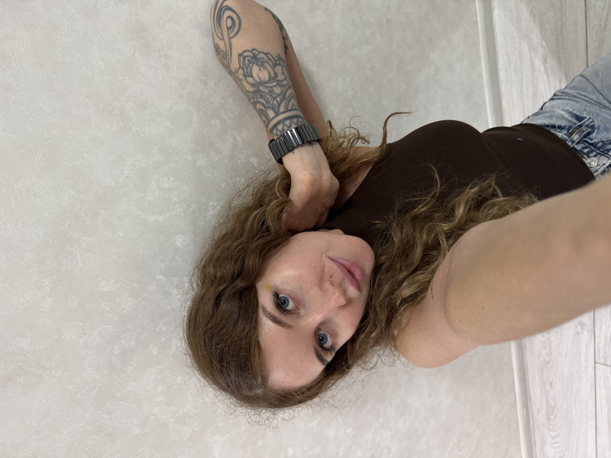 A young adult female appears in a selfie taken indoors, within what appears to be a residential interior in Chaikovsky, Russia. She has long, wavy brown hair, blue eyes, and a small stud piercing on her right nostril. A subtle yellow discoloration is visible above her right eyebrow. She wears a dark brown sleeveless top and light-wash denim jeans, partially visible at the bottom. On her left wrist, she wears a black, rectangular-linked bracelet. Her left forearm and upper arm are adorned with a prominent black and grey floral/ornamental tattoo. Her left hand is positioned behind her head, slightly touching her hair, as she looks directly at the camera with a gentle smile. The background features a light beige or off-white textured wall. A white baseboard and light-toned wooden floorboards are visible at the bottom of the frame. The lighting is even and ambient, consistent with an indoor setting.