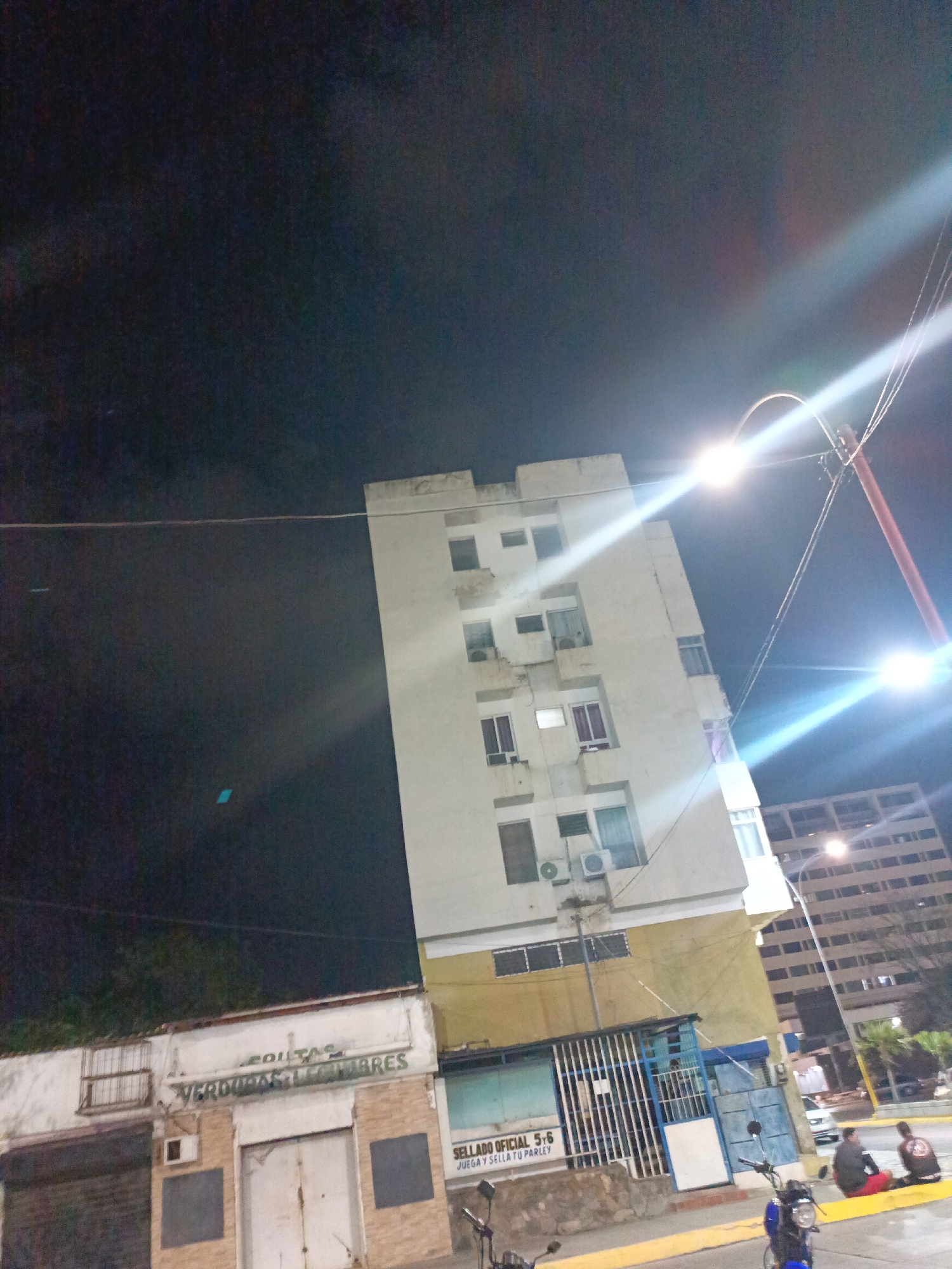 The image depicts a street scene in Juan Griego, Venezuela, at night. A tall, light-colored building dominates the frame, featuring multiple stories and windows, some with visible air conditioning units. Below the building is a series of small storefronts with closed metal gates, indicating they are likely closed for the night. One storefront is labeled "FRUTAS VERDURAS LEGUMBRES," suggesting it is a fruit and vegetable market. A sign on another store reads "SELLADO OFICIAL 5-6 JUEGA Y SELLA TU PARLEY." A street light is positioned in front of the tall building, emitting a bright glare. In the lower right corner of the image, individuals are sitting on the sidewalk, with parked motorcycles nearby. Another large building is seen in the background. The sky is dark, suggesting a late hour.