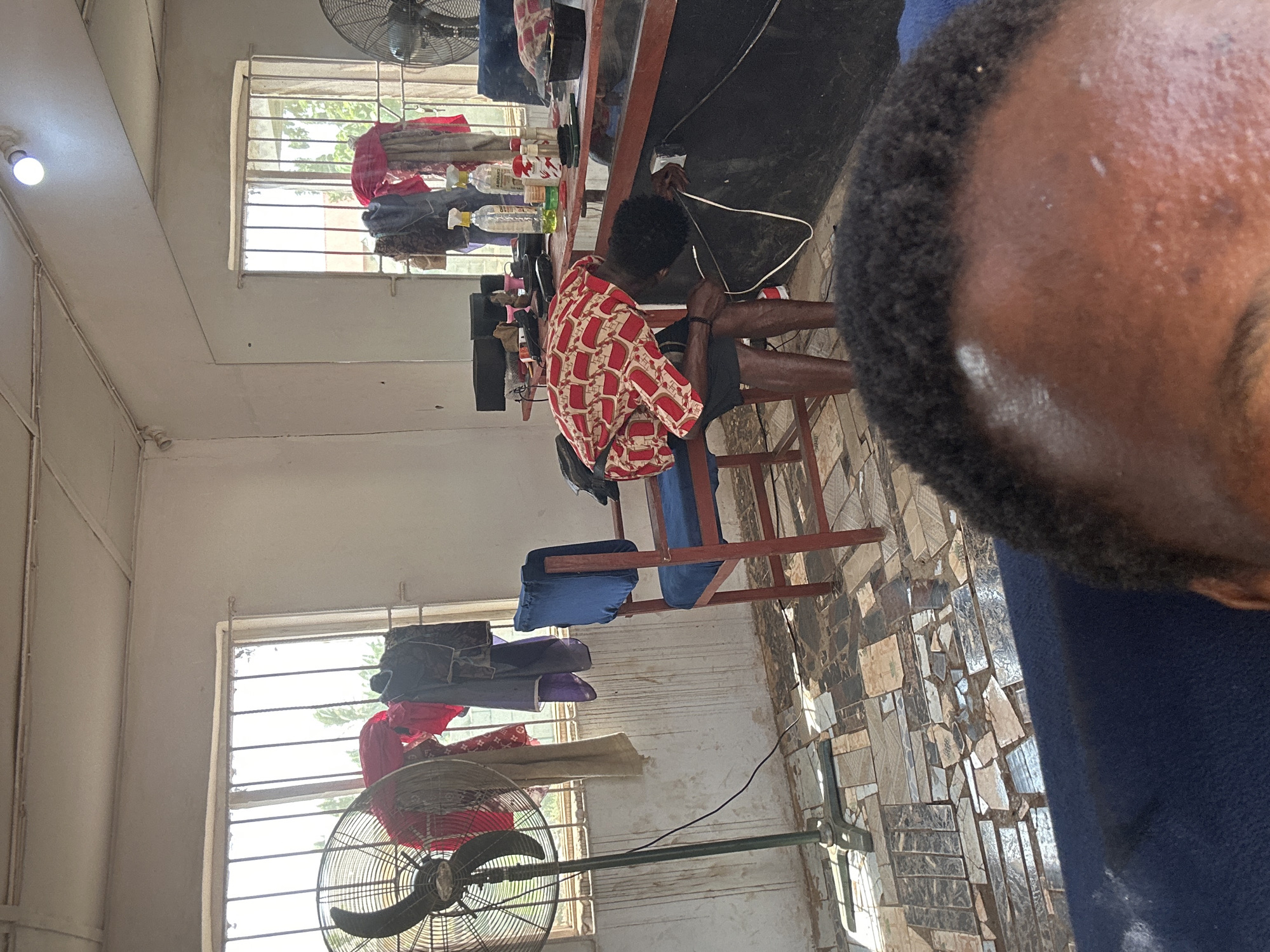 The image depicts the interior of what appears to be a barber shop. The location is in the city of Unknown, Unknown. A man in a red and white patterned shirt is sitting at a counter, potentially tending to electrical cords. The counter holds an assortment of bottles and barber tools. In the foreground, part of a person's head is visible, suggesting they are holding the camera or are very close to the subject. Two windows with metal bars are present, one near the top and one near the bottom of the frame, each with a fan positioned in front. Garments are hanging near the windows. The floor has a patterned tiling. Chairs with blue cushions are placed in the center of the room.
