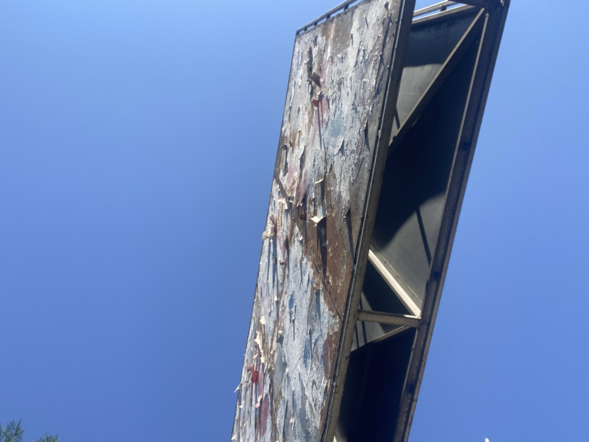 An outdoor, low-angle shot captures a damaged billboard against a clear blue sky. The billboard, positioned at an angle, shows significant wear and tear, with large sections of paint peeling off, revealing rust and underlying material. The structure supporting the billboard is made of metal beams. The scene is devoid of people or any discernible activity. It is likely situated in the city of Chornomorsk, Ukraine, given the context.