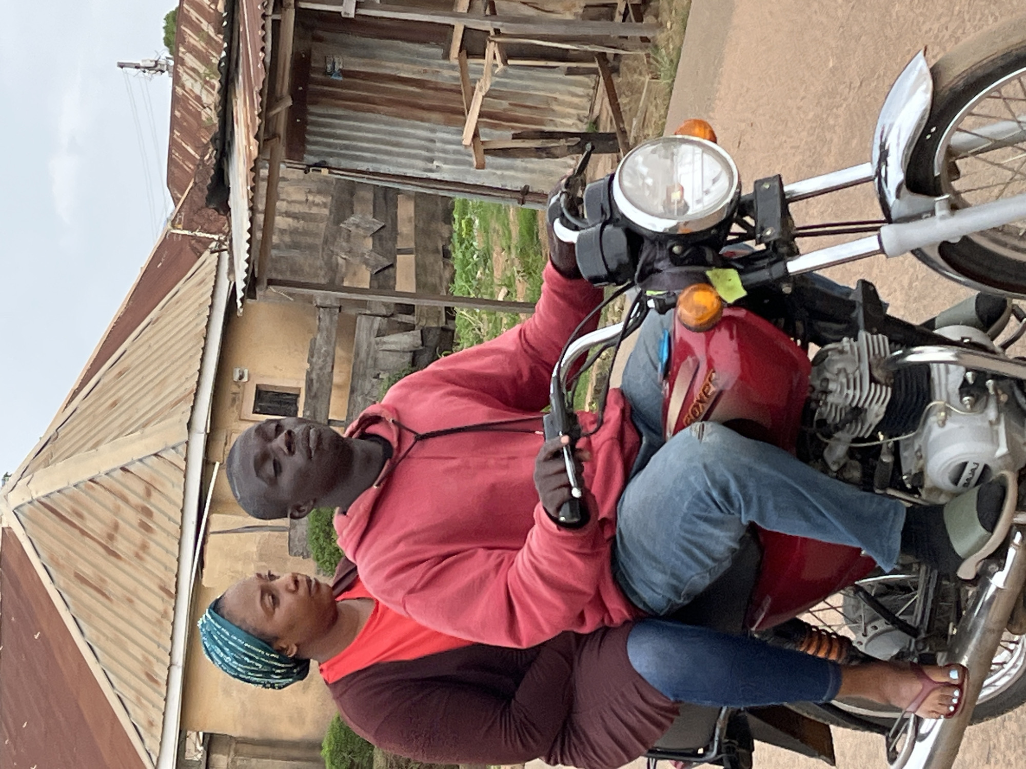 A man and a woman are seated on a red motorcycle on a paved surface, possibly a road. The man is operating the motorcycle, holding the handlebar. He is wearing a red hoodie and blue jeans. The woman is sitting behind him, wearing a red and brown long-sleeved shirt, blue jeans, and a blue head covering.
The background includes buildings with corrugated metal roofs and wooden structures. Green vegetation is visible. This is the City of Jos, Nigeria.