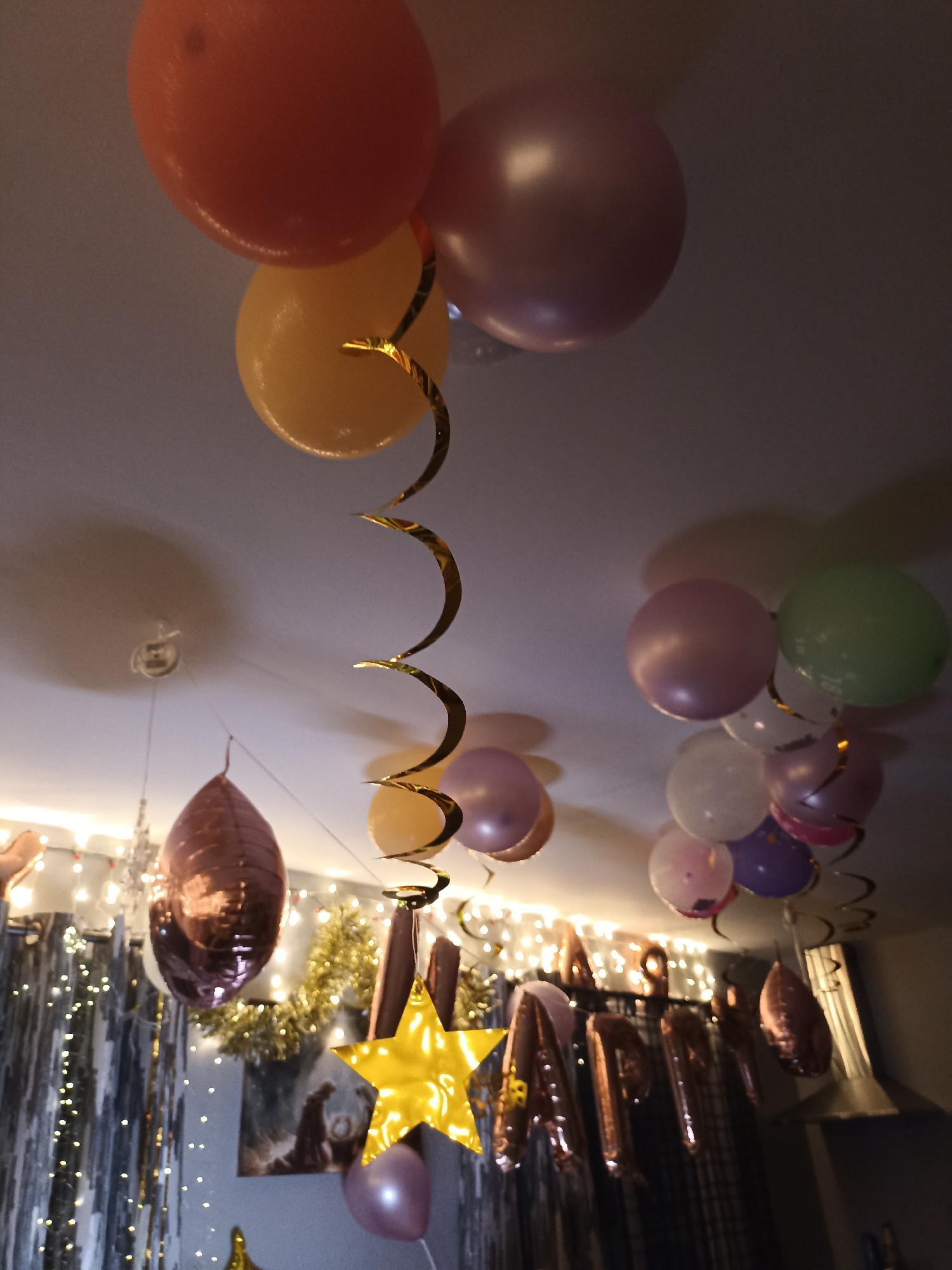 The image shows a ceiling decorated with balloons and streamers for a celebration. There are several balloons of various colors, including orange, yellow, pink, purple, and green, some of which are clustered together and others are hanging individually. Golden spiral streamers are also visible, adding to the festive atmosphere. In the lower part of the image, there are large foil letter balloons that appear to spell out "HAPPY BIRTHDAY", as well as a large gold star-shaped balloon. The background is dimly lit with fairy lights and metallic curtains. There are no people visible in the image. The scene is indoors, likely in a home or party venue. There is no information in the image that indicates the location is in Annagry, Ireland.