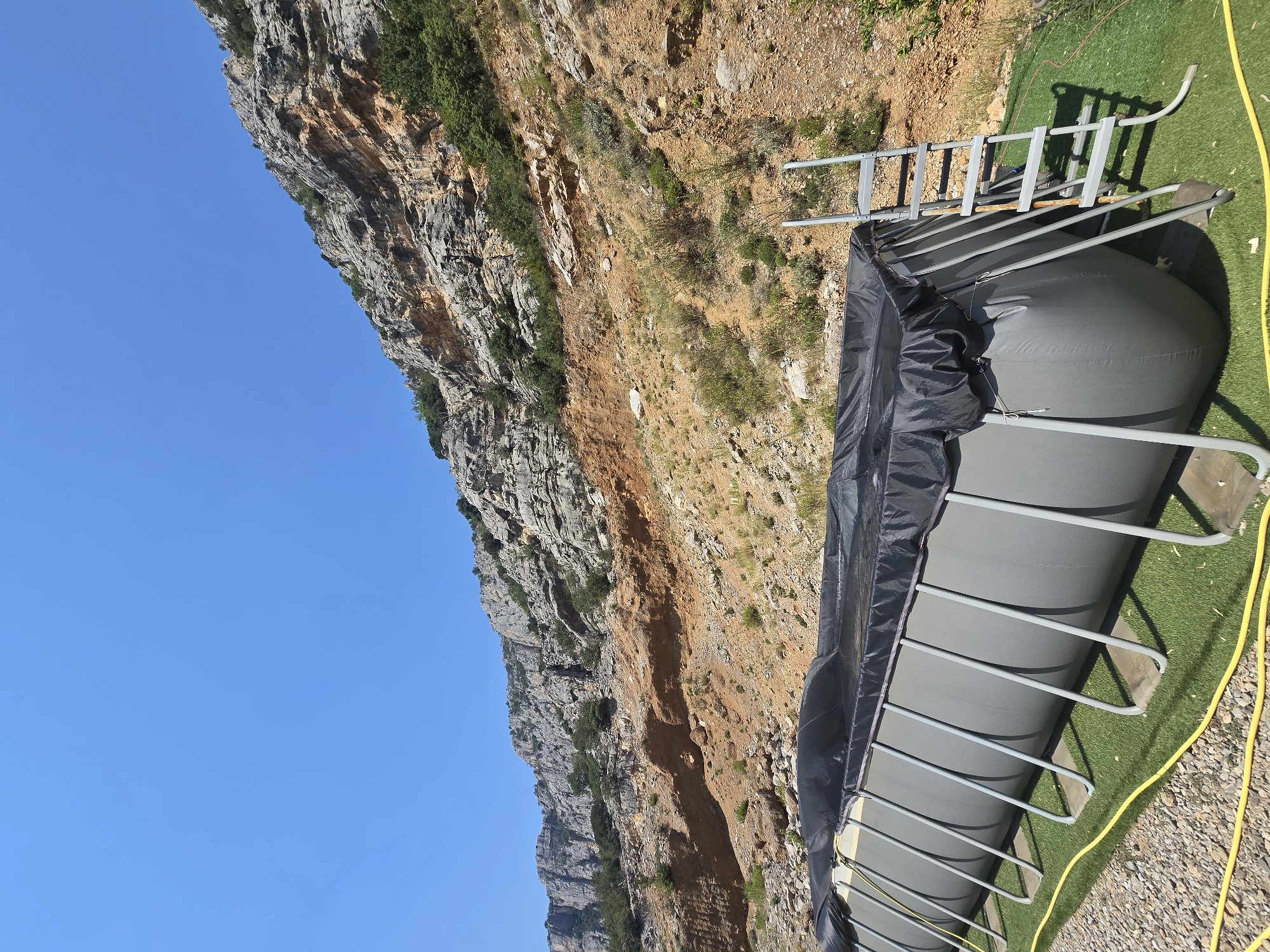 An outdoor, above-ground swimming pool is covered with a black tarp and a metal ladder rests against its side. The pool is situated on a patch of artificial grass, with a gravel area bordering it. In the background, a steep, rocky mountainside with sparse vegetation rises under a clear blue sky. The overall scene suggests a semi-rural or undeveloped location, possibly in the outskirts of Podgorica, Montenegro, given the characteristic mountainous terrain. No people are present in the image.