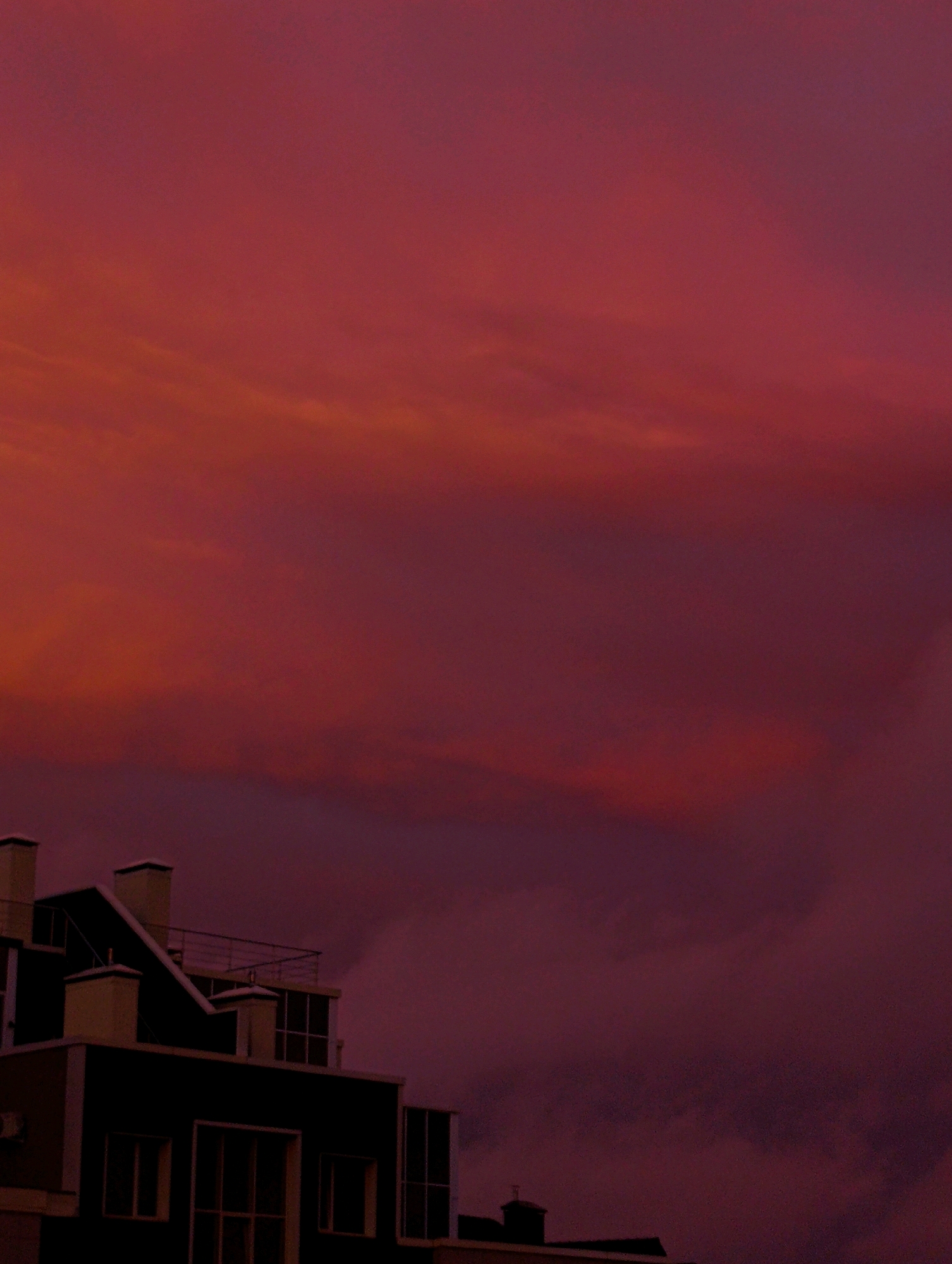 This is a medium shot, eye-level, exterior, natural light photograph of a modern building against a vividly colored sky. The sky dominates the frame with a gradient of deep reds and purples, suggesting either a sunset or a heavily filtered twilight. Wispy clouds are visible, blending into the intense colors of the sky. In the lower portion of the image, the top floors of a dark, contemporary building are visible. The architecture features sharp angles, large rectangular windows, and prominent, light-colored chimney-like structures. A metal railing lines a rooftop terrace. The scene is devoid of any people or animals. The overall atmosphere is dramatic and somewhat abstract due to the striking sky.