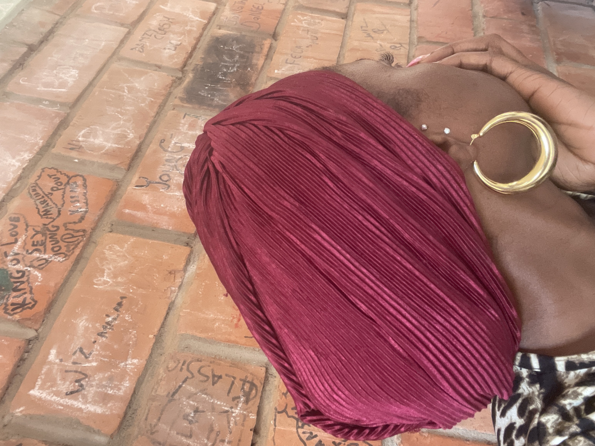 A close-up, profile view shows a person wearing a dark red turban and a gold hoop earring. The person's face is partially obscured by their hand, which is resting on their cheek. They are looking to the right, with visible eyelashes and two small studs in their ear. The background is a brick wall with white chalk graffiti. The graffiti is difficult to decipher in full, but some words appear to be "KING OF LOVE," "YOUNG," "CLASSIC," "WIZ," and "DANIEL." The scene is outdoors, possibly in a public space like a street or courtyard, given the graffiti. The lighting suggests it is daytime. There is no indication of Television, Nigeria in the image.