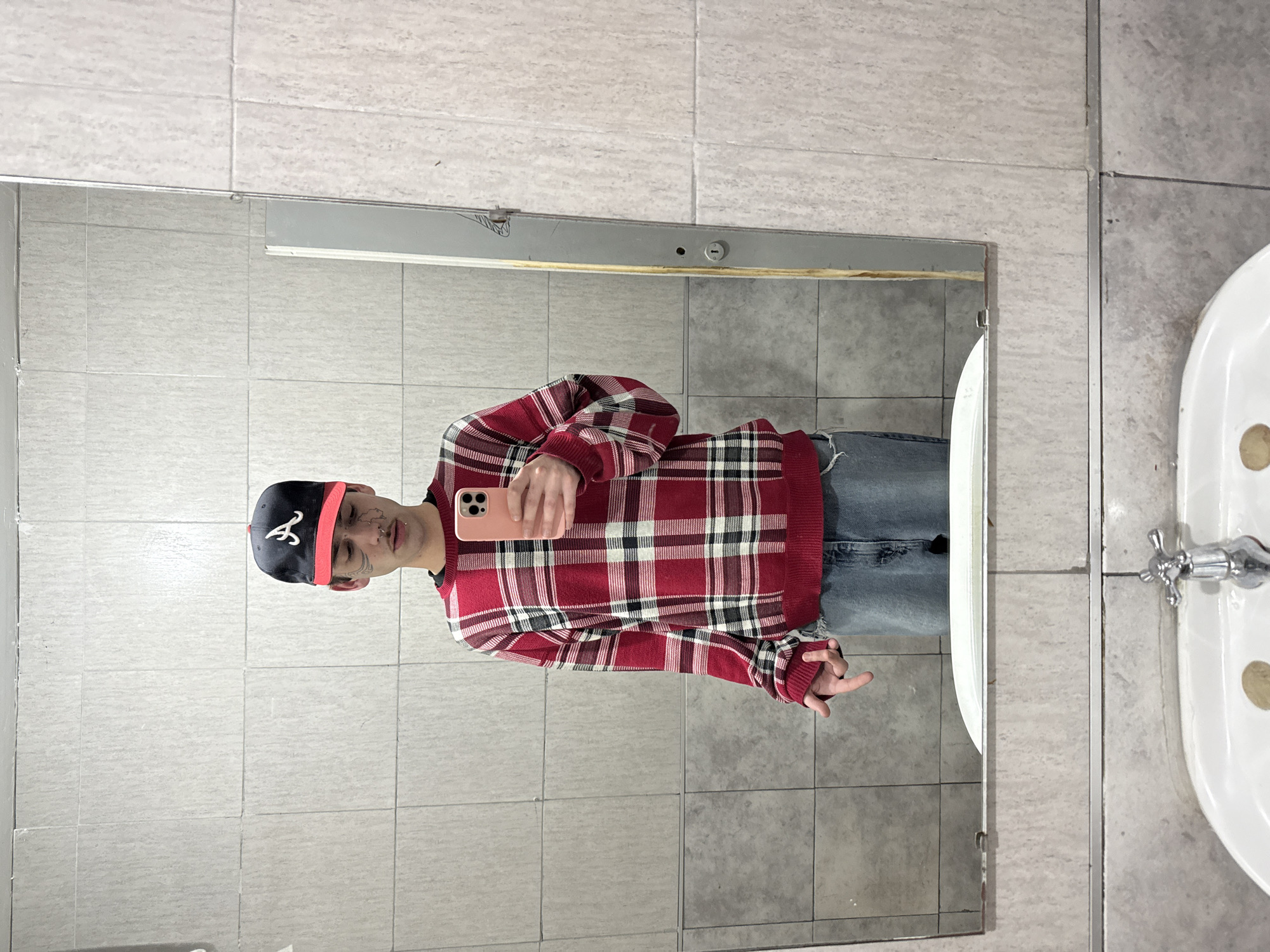 A person stands in a bathroom in what appears to be a public restroom, taking a selfie in the mirror. The person is wearing a red plaid sweater, blue jeans, and a baseball cap with a red brim. The bathroom has tiled walls and floors, a sink, and a mirror. The location in the city of Buenos Aires, Argentina cannot be confirmed based on the image.