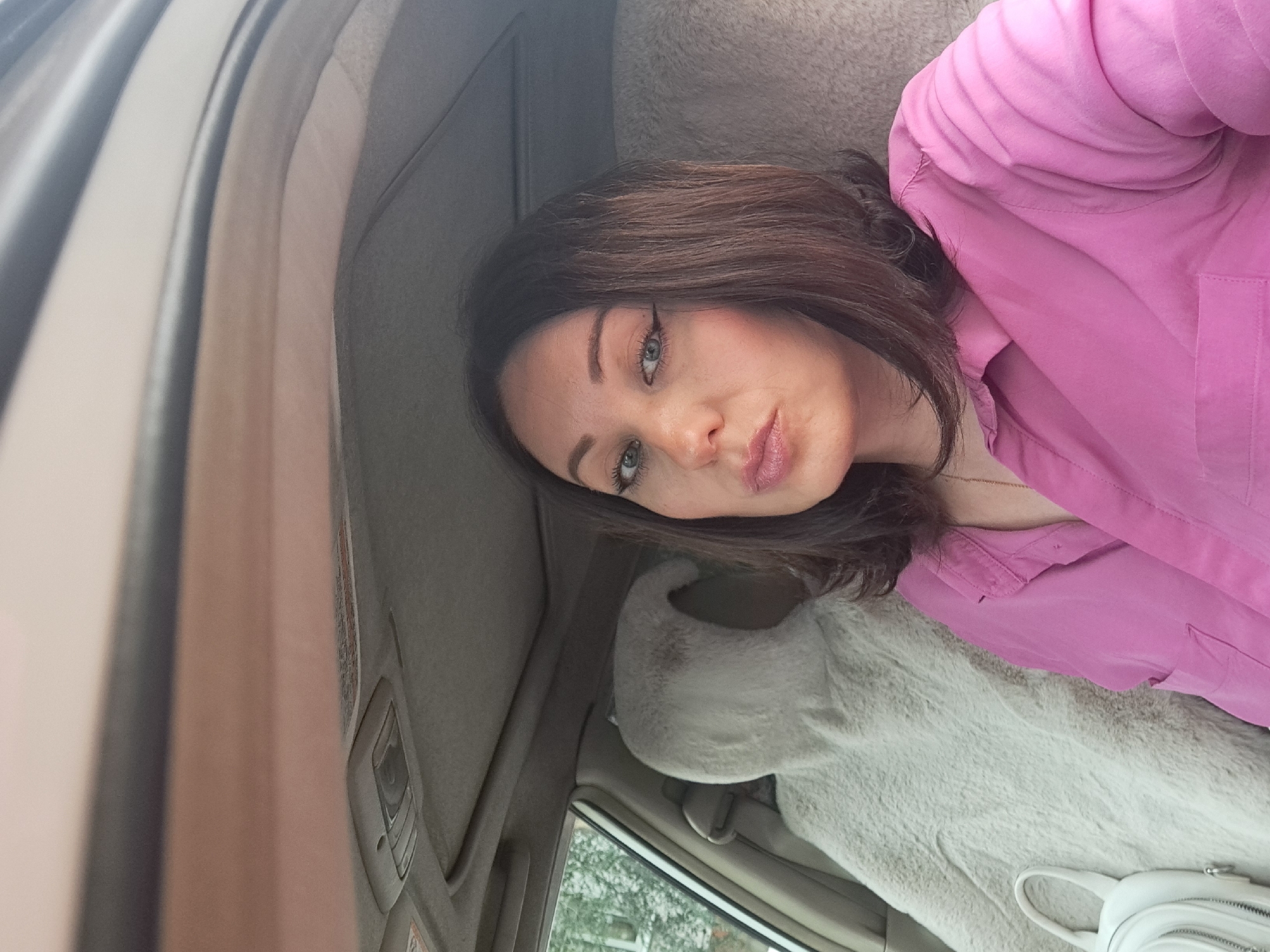 A woman with shoulder-length brown hair and winged eyeliner is sitting in the passenger seat of a car. She is wearing a pink collared shirt. The car's interior is visible, including a fuzzy seat cover on the passenger seat next to her and the car's ceiling with a sunroof. Outside the car window, trees and foliage are visible, suggesting a natural environment. The image appears to be a selfie, taken by the woman. There is no specific information in the image that indicates the location is Irkutsk, Russia.
