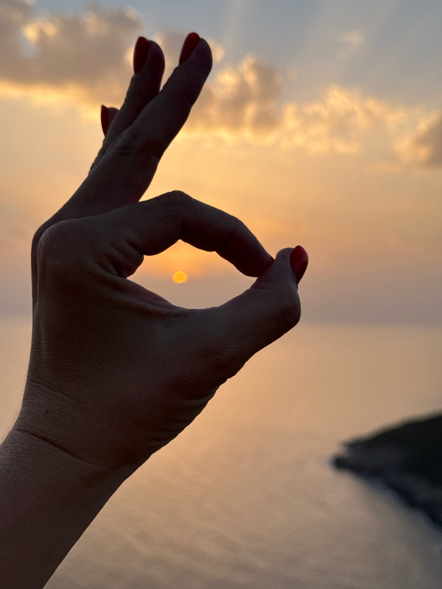 A hand forms an "okay" gesture against a hazy sunset sky, framing the setting sun. The fingers are silhouetted, with red nail polish visible on the tips. The background transitions from a warm orange glow near the horizon to a muted blue and grey sky. The water below is a soft, pale orange, with a distant dark landmass on the right. The scene evokes a sense of peace and appreciation for nature's beauty at dusk. The location is the city of Zagora, Montenegro.