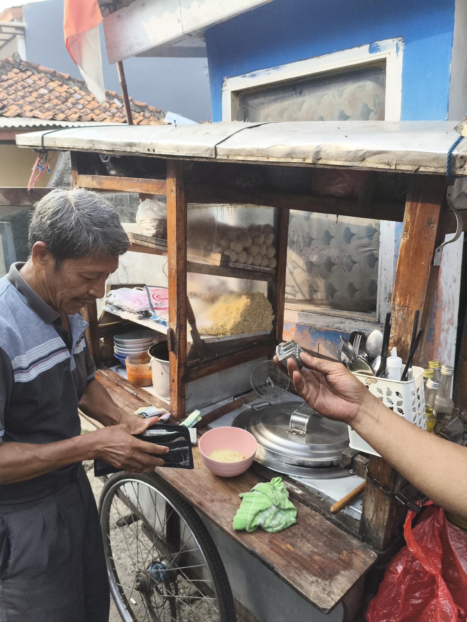 An outdoor food stall in Depok, Indonesia, shows a man with graying hair and a blue and gray striped shirt holding an open black wallet. He is standing to the left of a wooden food cart, which displays various food items and cooking utensils. In front of him on the cart is a pink bowl with food, a green cloth, and a bicycle wheel. A person's arm and hand are visible on the right, holding a piece of metal with a pattern. The background shows a building with blue and white paint, tiled roofs, and an Indonesian flag. The scene appears to be a transaction taking place at a street food vendor.