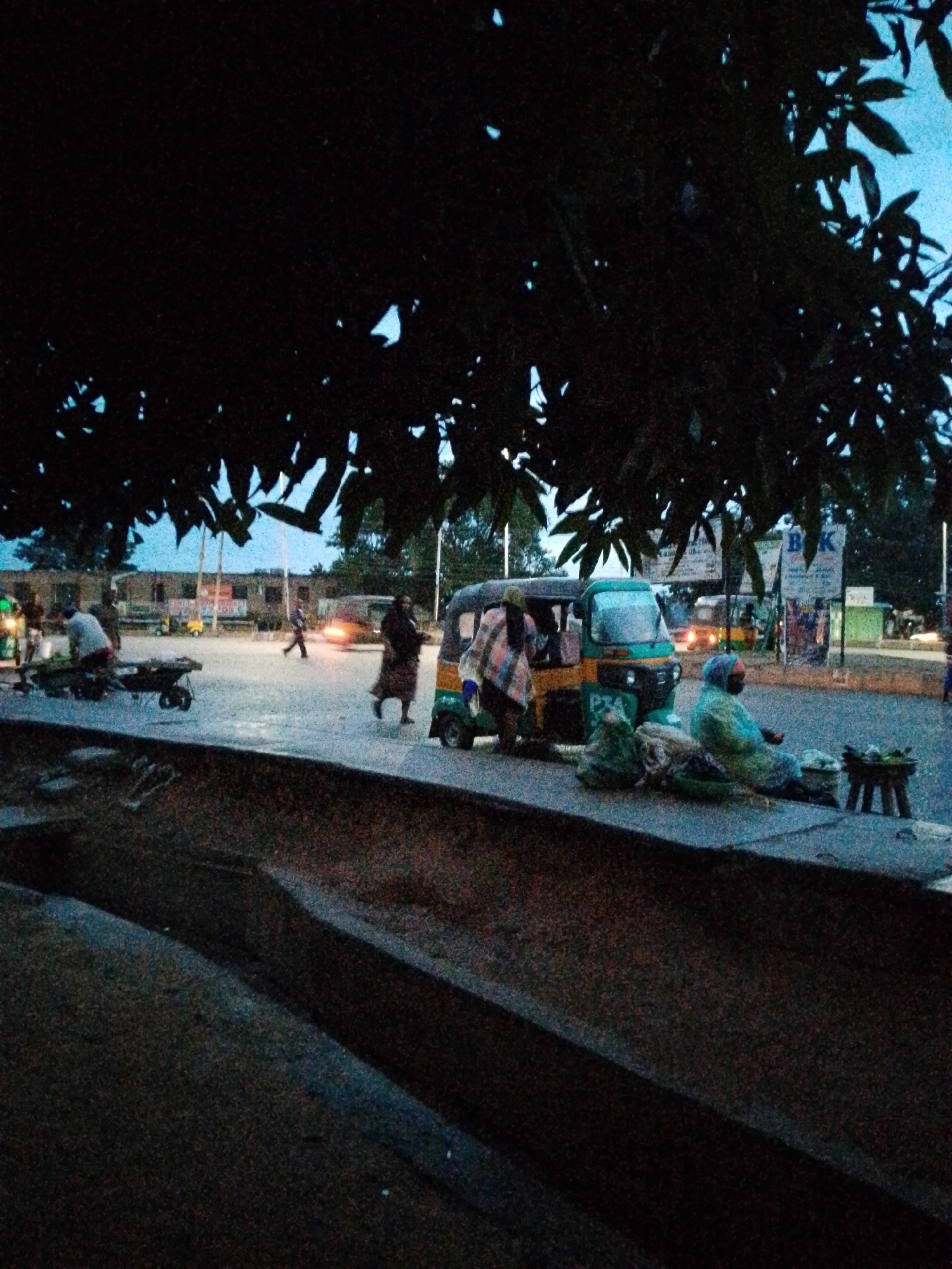 The image depicts a twilight street scene in Bukuru, Nigeria, likely at dusk or dawn, characterized by low light and a blueish hue in the sky. Dominating the upper foreground are large, dark trees, their foliage silhouetted against the ambient light.
In the mid-ground, a paved road or market area shows ongoing activity. A prominent green and yellow three-wheeled auto-rickshaw, common in the region, is parked on the right side of the road, with the faded letters "PTA" visible on its front side panel. Nearby, two women are seated on the ground, engaging in what appears to be roadside commerce. One woman, clad in a light blue/green garment, tends to items displayed on a small wooden stool, with bags and bundles surrounding her. The other woman, partially visible, is draped in a patterned blanket or shawl. Another person stands beside the auto-rickshaw.
Further left, a figure pushes a cart or wheelbarrow, and a lone individual walks across the street. In the background, the soft glow of headlights from another vehicle adds an orange tint to the scene. Buildings and what appear to be street signs or billboards are discernible, with "BK" clearly visible on one sign, followed by partially obscured text "MA... B...". The ground in the immediate foreground consists of a dark concrete structure or curb. The scene suggests a bustling marketplace or street during the transitional hours, with daily activities continuing under dim natural and artificial light.