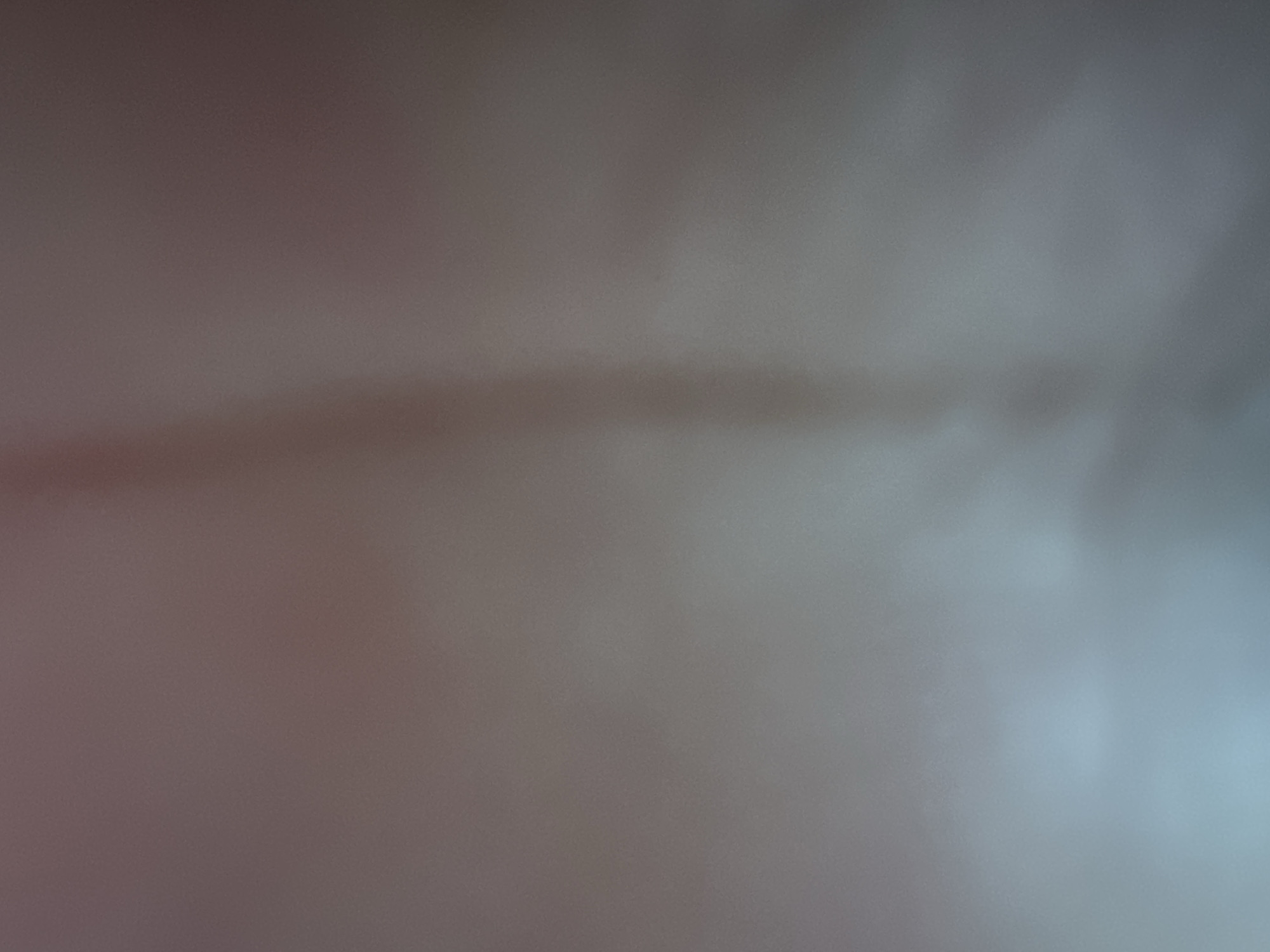 The image is an abstract and blurry representation of a scene, likely taken indoors or under artificial lighting, given the soft focus and muted color palette. There are no discernible subjects like people or distinct objects. The overall impression is of a textured surface with subtle variations in light and shadow. A horizontal band of darker, elongated blur stretches across the middle of the frame, suggesting a shadow or a directional light source. The left side of the image is dominated by warm, reddish-brown tones, while the right side transitions into cooler, bluish-grey hues. This gradient of color, combined with the lack of sharp detail, creates a sense of depth and atmosphere, but the exact nature of the scene remains ambiguous. There is no visible text. Given the provided location context, it is impossible to infer any specific details about the setting or event from the image itself.