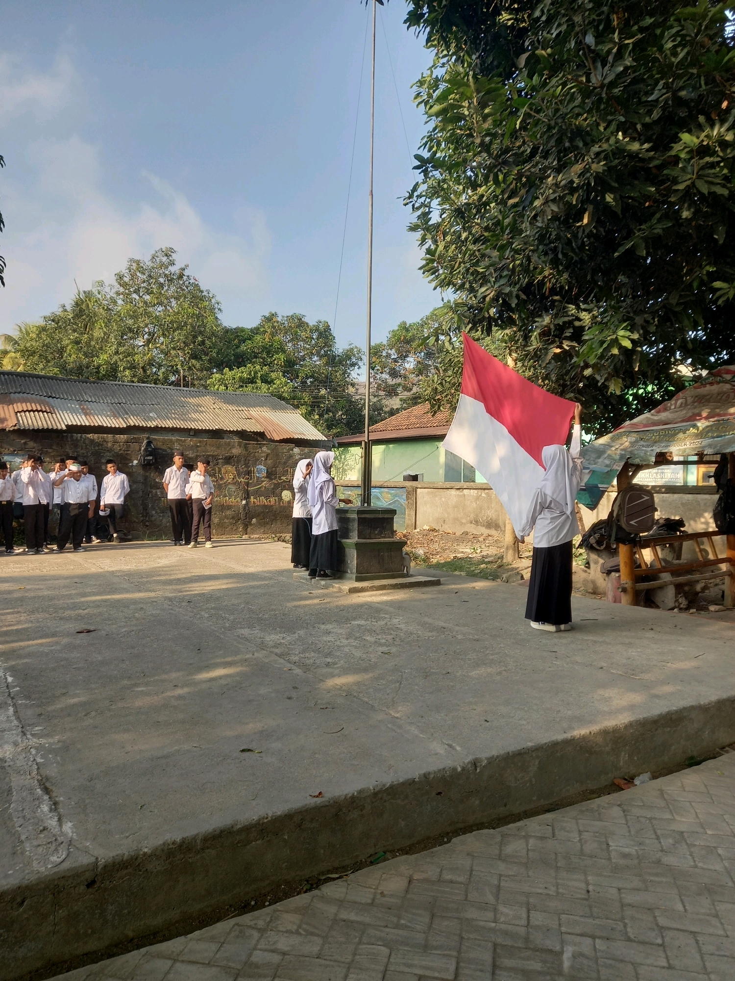 The image depicts an outdoor flag-raising ceremony at what appears to be a school in the village of Perampuan, Indonesia. A group of students, dressed in white shirts and black pants or skirts, stand in formation, some saluting. Two female students, one holding a red and white Indonesian flag, are positioned near a flagpole. The background includes a weathered building with a corrugated metal roof, a brick wall with graffiti, and lush green trees. The ground is paved with concrete. The sky is clear with some light clouds.