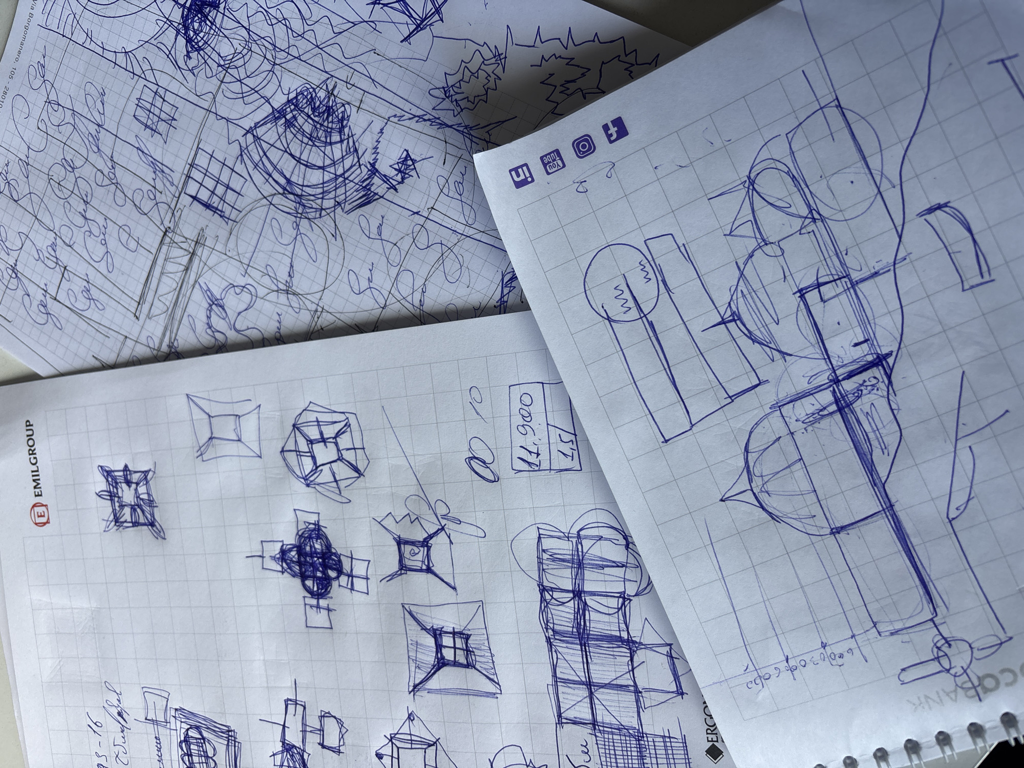 This overhead shot captures several overlapping sheets of paper, likely from notebooks, laid out on a light surface, suggesting a creative or planning session taking place in Yerevan, Armenia.
All papers are gridded with blue lines and are covered in intricate sketches and handwritten notes made with blue ink. The paper at the top-left displays dense, abstract linear drawings, grid patterns, and free-flowing squiggles, possibly representing architectural, mechanical, or conceptual designs.
A sheet in the middle-left features numerous geometric designs, primarily square and cubic forms rendered in various perspectives, some containing internal structures or radiating lines. This sheet prominently displays the "E EMILGROUP" logo along its left edge. Numerical figures such as "11,900" and "1,51" are visible within a box, alongside other handwritten numbers and letters like "93-16" and "0010".
The paper on the right, which is spiral-bound at the bottom, showcases more complex, multi-layered sketches involving prominent circles, cylinders, and interwoven lines, hinting at abstract or technical concepts. This sheet also has social media icons for LinkedIn, YouTube, Instagram, and Facebook printed upside down along its top edge. Partially visible text near the binding includes "ERGO" and "COBANK". Some faint handwritten notes and calculations, including an address "Via Borgomanero, 105 - 280", are interspersed throughout the drawings on all papers. The bright, even lighting suggests an indoor setting.