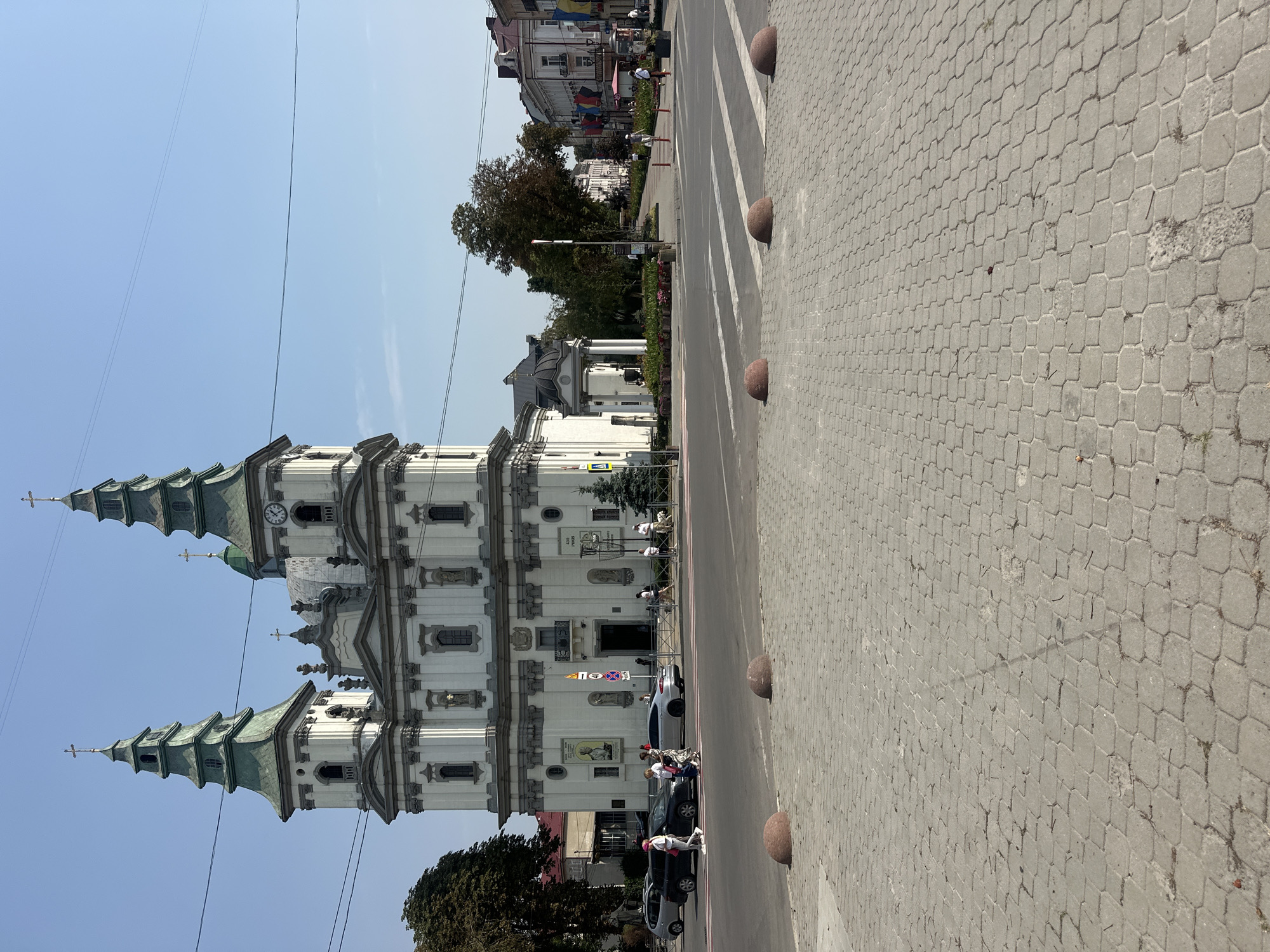 The image captures a street view of the Ternopil City Hall in Ternopil, Ukraine, on a clear day. The Ternopil City Hall, a prominent white building with two green-capped towers topped with crosses, dominates the background. The building features classical architectural elements with ornate decorations. A clock is visible on one of the towers.
In the foreground, a pedestrian crossing is marked with white stripes on a paved road. Red, rounded bollards are strategically placed along the edge of the pedestrian area. The pavement itself is made of interlocking hexagonal concrete pavers, with some areas showing signs of wear and grass growing through cracks.
Several people are visible in the scene. On the pedestrian crossing, a group of individuals are walking, including a person wearing a pink helmet. Cars are also present, including a black sedan and a silver sedan. On the right side of the street, more pedestrians can be seen walking, and a dog on a leash is accompanied by its owner.
The environment includes trees, some with full foliage and one with sparser leaves, indicating a likely late summer or early autumn setting. Buildings line the street in the mid-ground and background, some displaying colorful flags. Overhead, power lines stretch across the frame. The sky is a clear, bright blue. The overall scene depicts a normal day in a city square, with the grand architecture of the City Hall serving as a focal point.