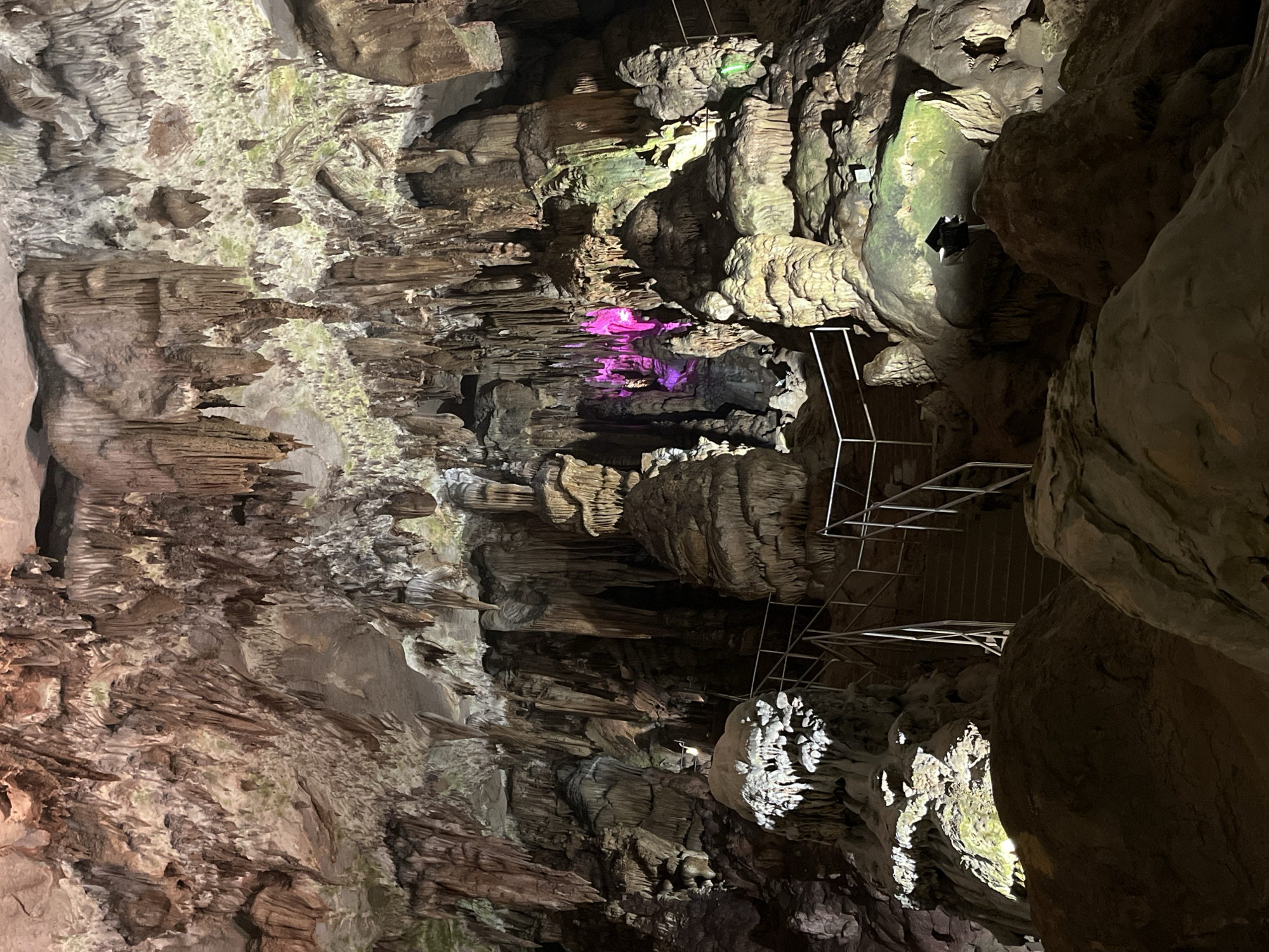 The image depicts the interior of a cave, likely a tourist attraction given the presence of a metal railing and steps. The cave is adorned with numerous stalactites hanging from the ceiling and stalagmites rising from the floor, creating a complex and textured environment. The rock formations display a variety of colors, including earthy browns, grays, and whites, with some areas illuminated by artificial lighting that casts colorful hues of pink and green onto the formations. A set of stairs with a handrail is visible, leading upwards into the cave. The overall scene is one of natural geological wonder enhanced by man-made structures for accessibility.