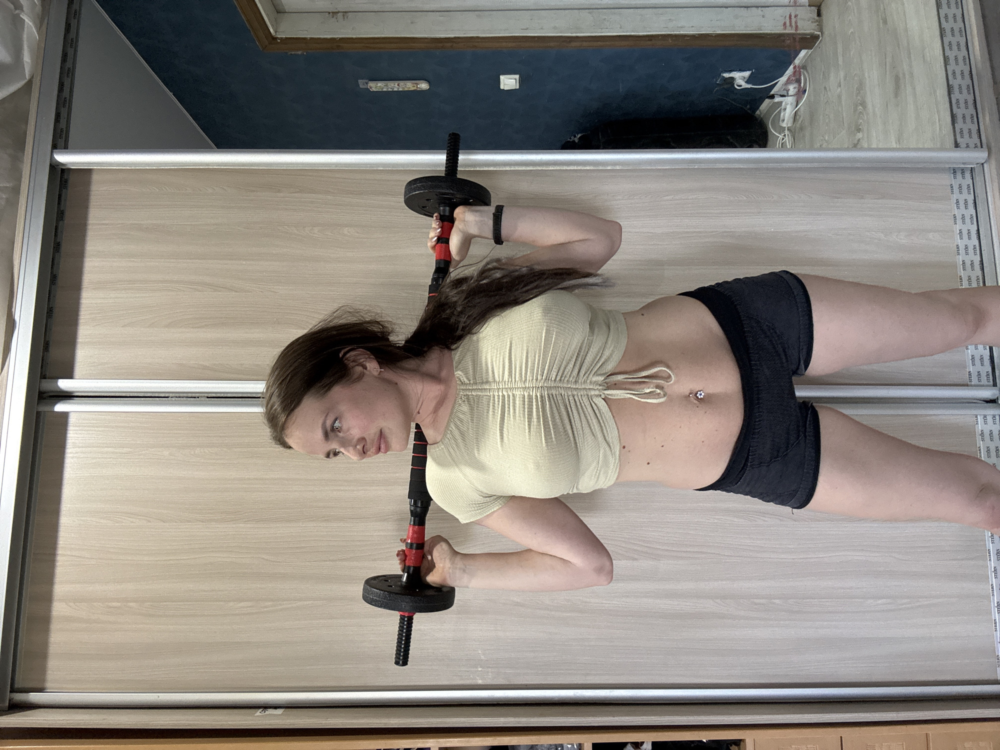A woman is holding a barbell in front of a closet. She is wearing a light green crop top and black shorts. She has a belly button ring. The closet doors are made of wood. In the background, there is a blue wall and a floor with wooden planks. A white electrical cord and plug are visible on the blue wall.