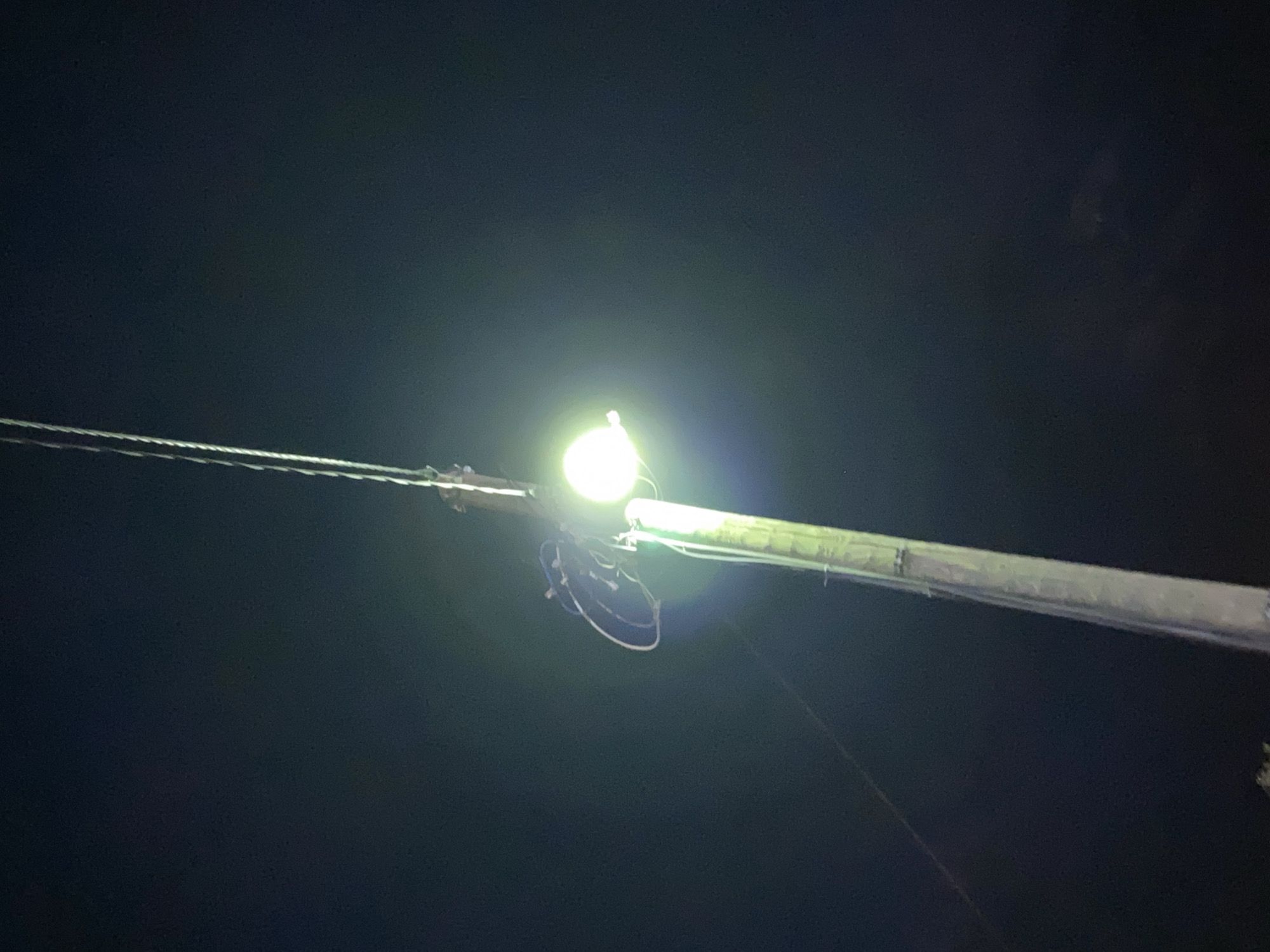 A pair of shoes is suspended from a utility pole near a street light. The scene is at night and it is dark. The location is unknown.