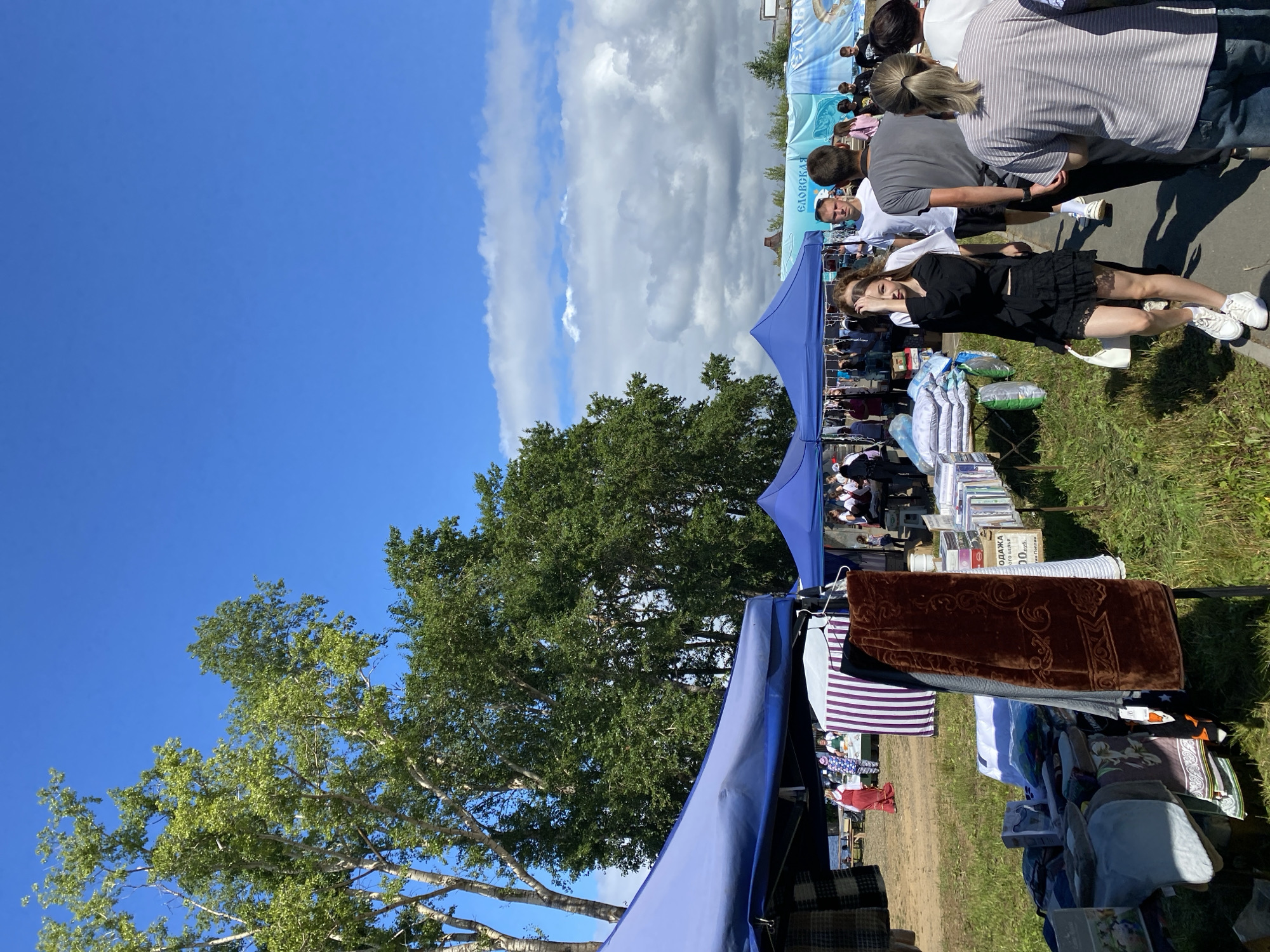 An outdoor market is depicted under a bright blue sky with scattered clouds. In the foreground, a young woman in a black skirt and top walks towards the viewer, carrying a white handbag. To her left, a stall displays textiles, including a dark brown velvet cloth with an embroidered pattern. Further back, several tents house various goods and people are browsing. A banner with Russian text, including "ЕЛОВСКАЯ", is visible in the background. The ground is a mix of dirt and grass, with a paved pathway in the foreground. Trees with green foliage are prominent on the left side of the image.