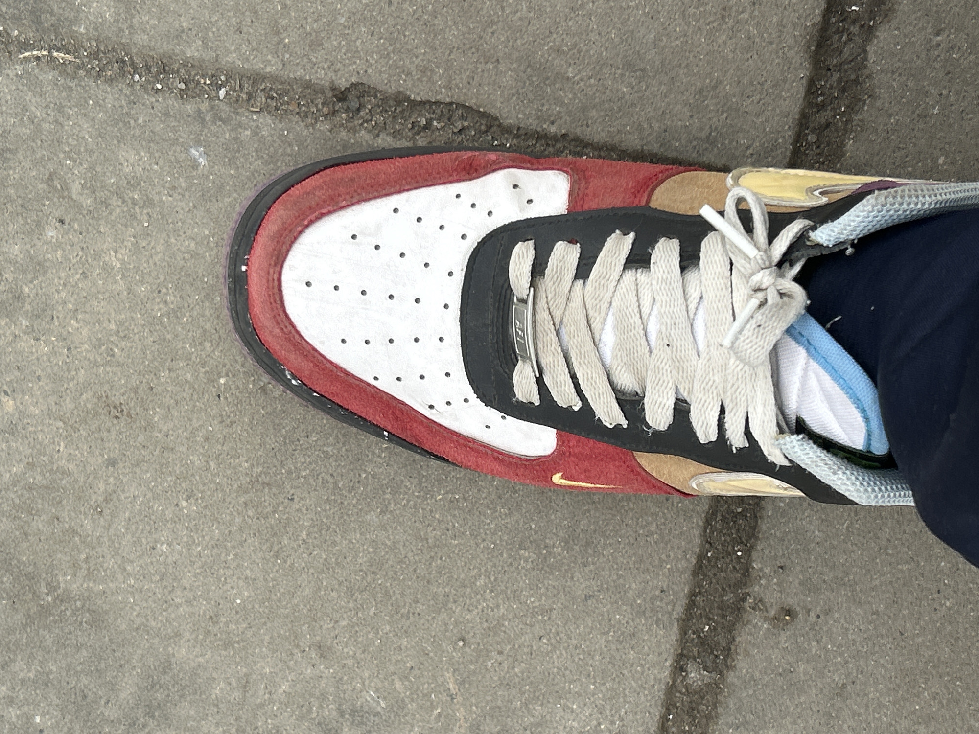 The image shows a single Nike Air Force 1 shoe on an asphalt surface. The shoe has a red suede border around a white perforated leather toe box. The area around the laces is black, and the laces are light grey. A small gold Nike swoosh is visible on the side. A dark blue fabric is visible near the ankle of the shoe, likely clothing. The asphalt has a visible crack. The location appears to be outdoors on a street or sidewalk. There is no indication of the location being Burgas, Bulgaria in the image.