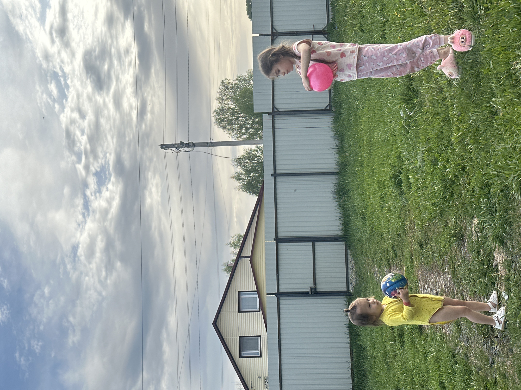 The image shows two young girls in a grassy yard near a house and a fence. The sky is cloudy. One girl is wearing pink pajamas and holding a pink ball. The other girl is wearing a yellow outfit and holding a multi-colored ball. The location is likely Kharnavy, Russia, based on the context provided in the prompt. Utility poles with power lines are visible in the background.