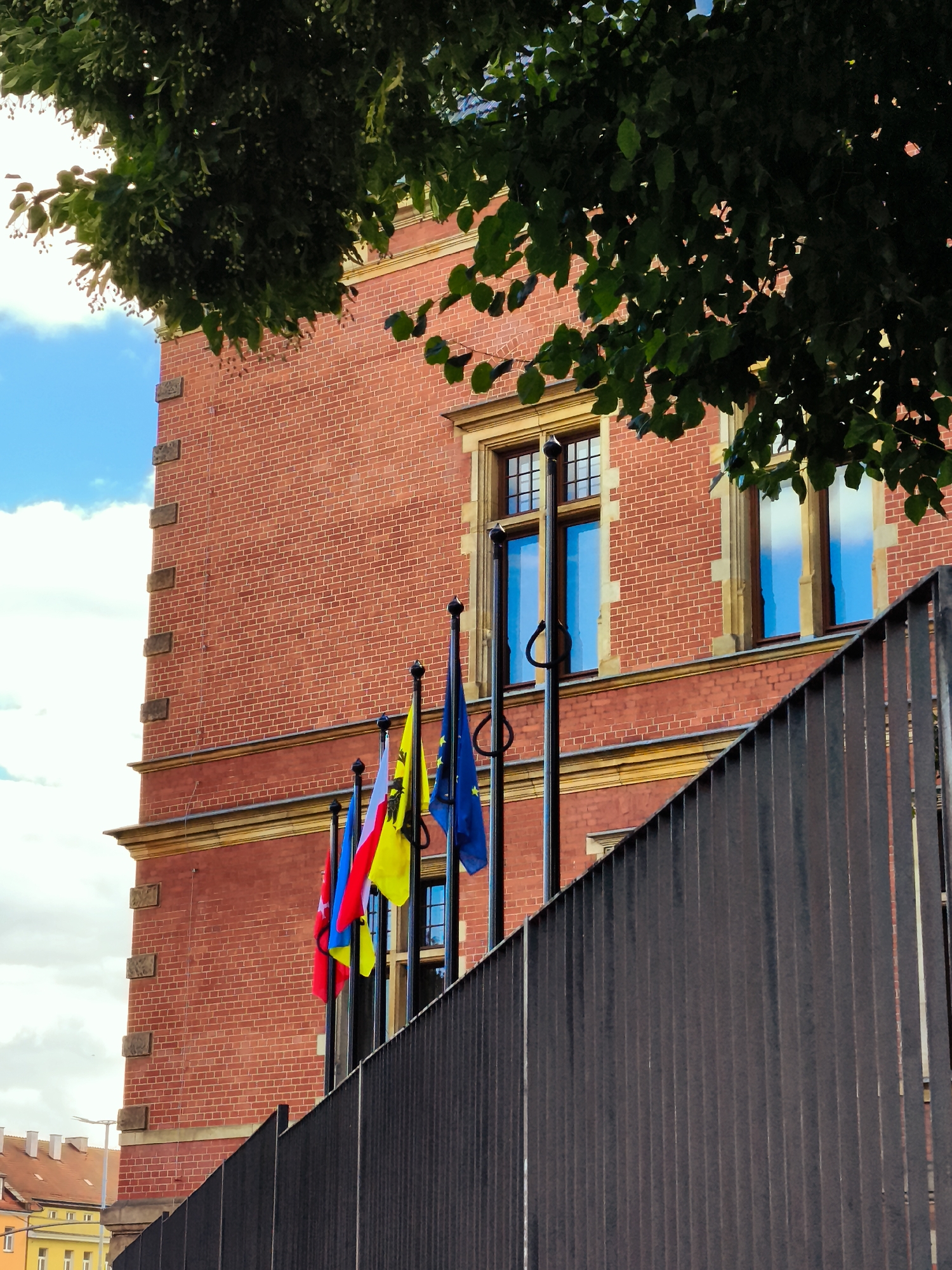 The image shows the facade of a red brick building with yellow decorative elements and windows. Several flags are displayed on poles in front of the building. In the foreground, a dark, slatted fence runs across the frame. The sky above is blue with scattered clouds, and a portion of a tree with green leaves is visible in the upper left corner. The building's architecture suggests a European setting, and the flags appear to be national and possibly European Union flags. The overall impression is one of a formal or institutional setting.
Based on visual cues, such as the architecture and the presence of flags that resemble the Polish and European Union flags, this scene is likely located in Gdańsk, Poland. Specifically, it is a view of the entrance to a significant public building, possibly a government office or historical landmark, in the city center. The dark fence in the foreground is a modern addition, likely for security or to delineate a public space from a restricted area. The sunlight filtering through the leaves of the tree suggests it is daytime, possibly late morning or early afternoon.