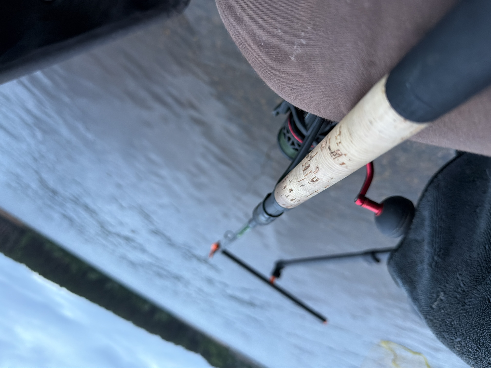 The image displays a close-up, low-angle perspective of an outdoor scene, situated in Chaikovsky, Russia, focusing on fishing activity. In the immediate foreground, the upper thighs of an individual, dressed in light brown or khaki trousers, are visible. A fishing rod, featuring a cork-wrapped handle and a black rubberized butt cap, rests horizontally across the individual's lap. A black spinning reel with a prominent red handle crank is mounted on the rod, partially visible beneath the rod handle. To the left, a textured dark grey fabric object is present. The fishing rod extends forward towards a large body of calm, dark blue-grey water, with its tip, equipped with an orange indicator, angled downwards. Further out on the water, a second black fishing rod or rod holder, also marked with orange tip indicators, is observed. The background reveals a distant, dark green treeline or forest forming the opposite shore of the body of water, beneath an overcast, light blue-grey sky.