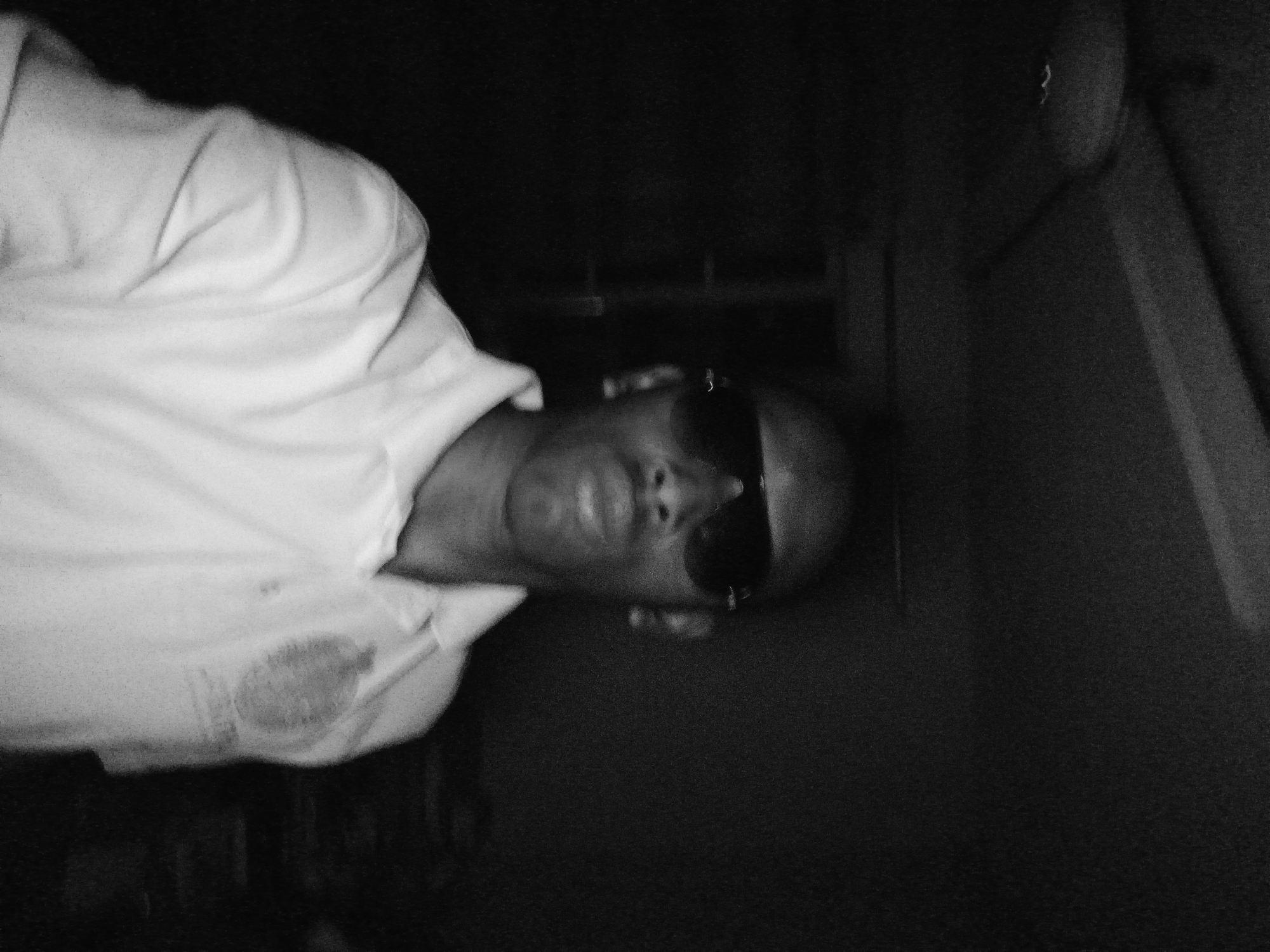 In a dimly lit interior, a person is captured in a low-resolution, black and white selfie. They are wearing sunglasses and a collared shirt, which appears to be white. The background is dark and indistinct, with what looks like a ceiling fan visible at the top of the frame. The overall image quality is poor, with significant grain and blur, making it difficult to discern further details about the subject or the surroundings. The location of Ikeja, Nigeria, is not discernible from the visual information present in the image.