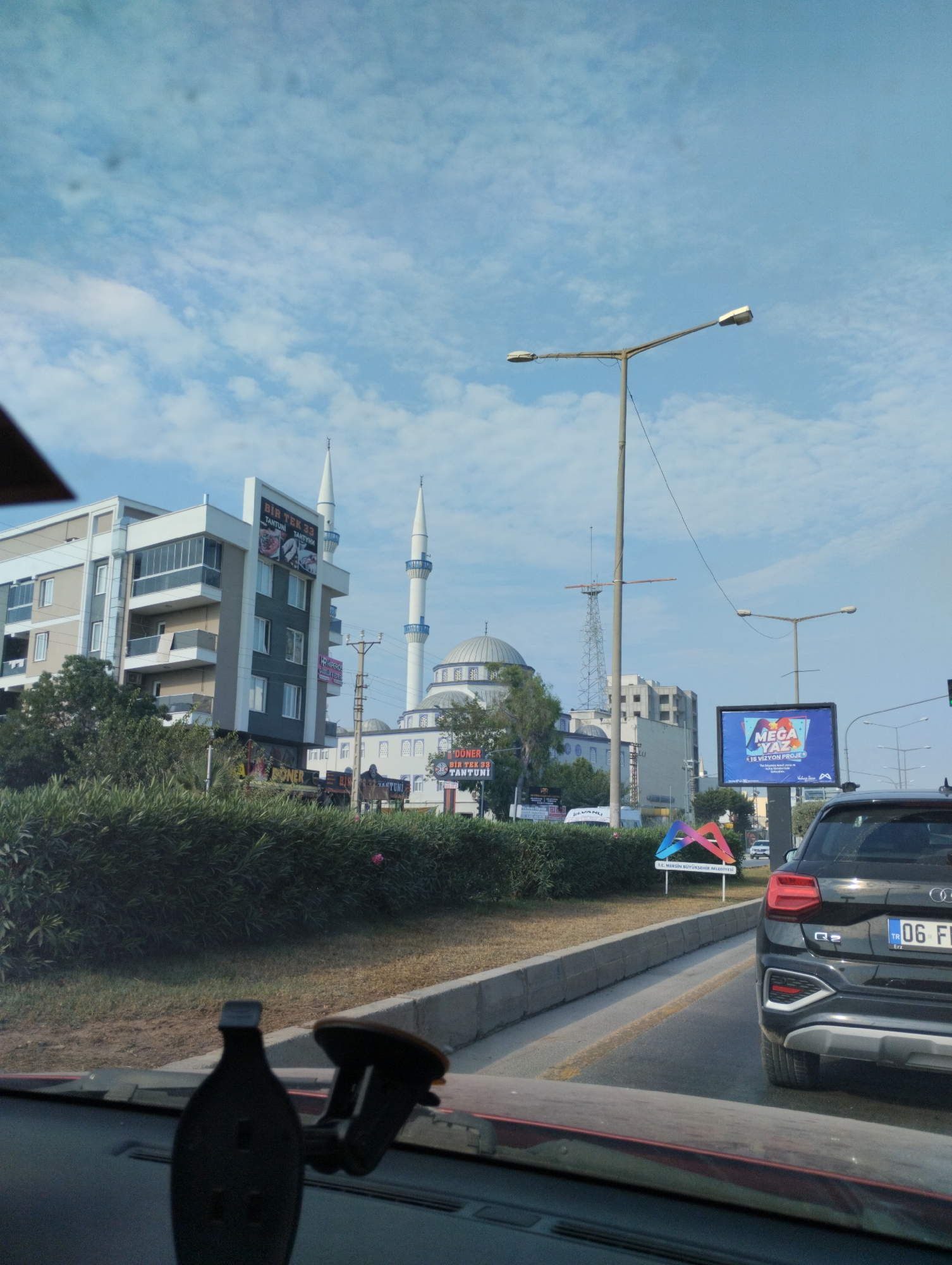 The image captures a street scene in Mezitli, Turkey, under a cloudy sky. In the foreground, the interior of a vehicle is visible, with a dashboard-mounted phone holder. The car is in motion, suggested by the blurred foreground elements.
The middle ground features a roadside with a dense green hedge lining the street. Beyond the hedge, a modern building with multiple floors and balconies stands prominently. To the right of this building, a mosque with a dome and two slender minarets is visible, characteristic of Turkish architecture. A large billboard advertising "MEGA YAZ" is also present, alongside streetlights and power lines.
In the background, other buildings and infrastructure are discernible, contributing to the urban environment. The overall scene depicts a typical day in a Turkish city, with a blend of residential, religious, and commercial elements. The presence of street signage in Turkish confirms the location.