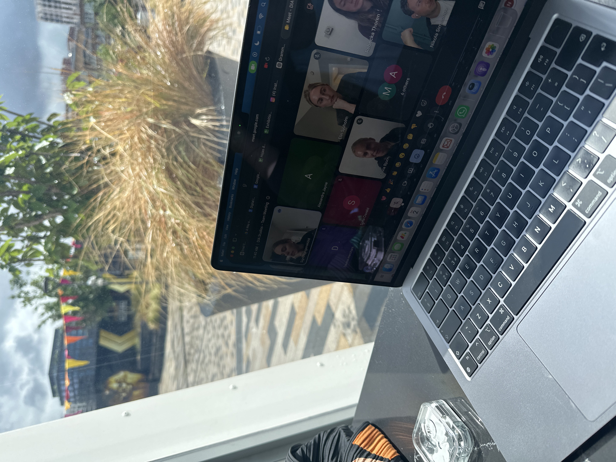 The image captures a close-up view of a silver MacBook laptop placed on a dark table next to a large window. The laptop screen displays an active Google Meet video conference titled "DIA Studios - Team Meeting," with the time "11:40 AM" visible. Multiple participants are seen in video frames, including Rebecka Thoren, Elin Sjöholm, Alexandra Jayne, Sofia Osvald, Olivia Bjorn, Nikola Simeonov, Sigrid Langenskiold, and Davina merchant, alongside others represented by initials (A, D, S, MA) and a "6 others" count. Browser tabs visible include "meet.google.com," "Email C...", "Digital I...", "Google...", "Gramm...", and "(4) Inst...". The menu bar shows "Safari File Edit View History Bookmarks Window Help".
Outside the window, a blurred urban landscape is visible. Prominent in the foreground are tall, dried ornamental grasses, with greener foliage and reflections of distant buildings, suggesting an outdoor garden or park area in a city like London, United Kingdom. One reflection shows a building with distinctive yellow and black chevron patterns and colorful triangular flags. The sky appears partly cloudy, and sunlight creates reflections and subtle shadows.
Next to the laptop, a clear, possibly glass or plastic, container is on the table. The laptop's keyboard, featuring a standard QWERTY layout with function keys (F1-F12), control, option, and command keys, is clearly visible. The overall scene depicts a remote work or study setup, likely in a cafe or office environment.