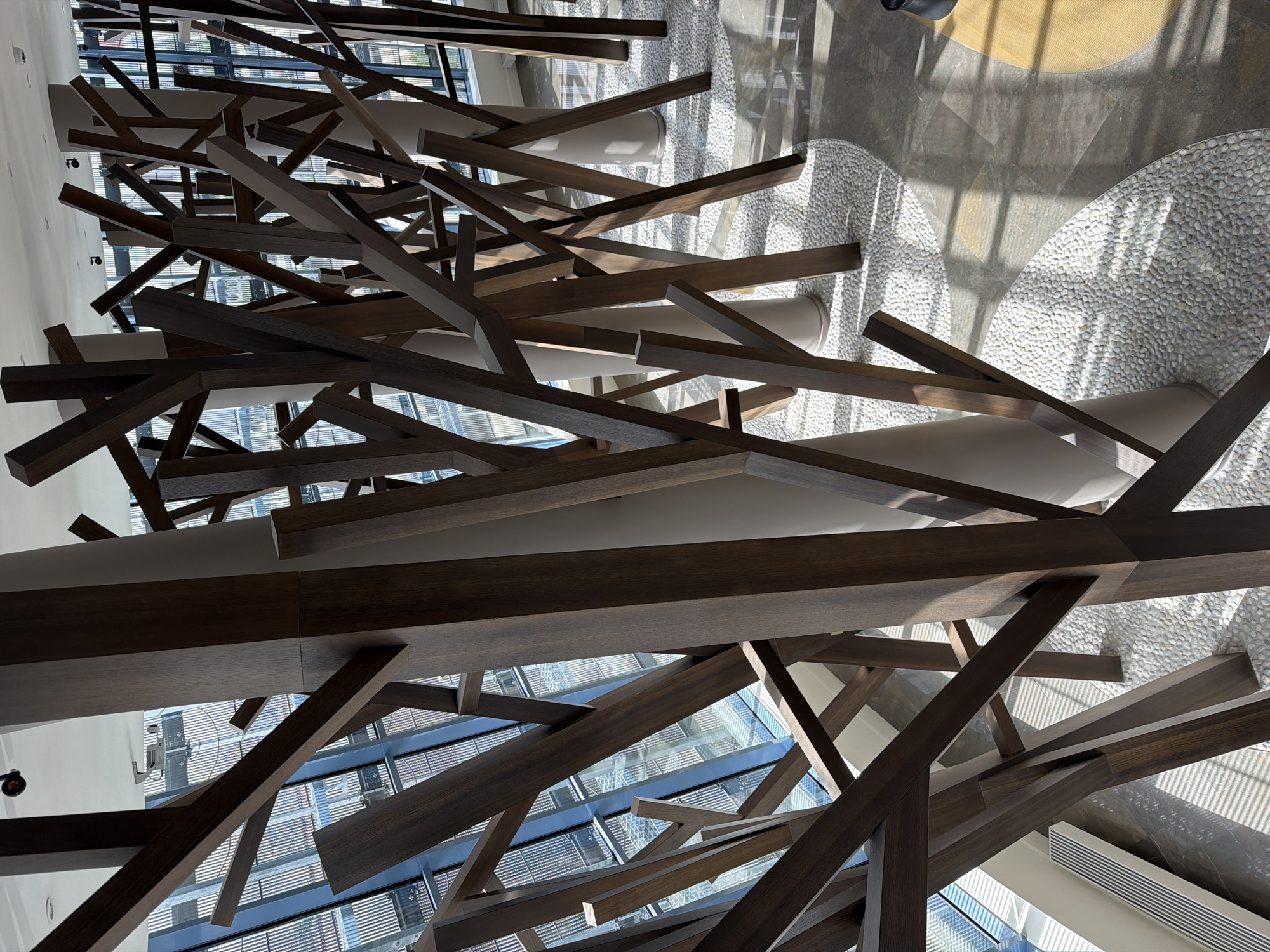 This image shows a modern architectural interior, likely a hotel lobby or atrium, featuring a complex, abstract installation of dark wood beams suspended from the ceiling. The beams are arranged at varying angles, creating a dynamic and visually striking pattern. Below, the floor is covered in a mosaic of small, light-colored stones, which contrast with the dark wood above. The presence of natural light filtering through windows, combined with the reflections on the polished floor, suggest a well-lit space. The scene captures a blend of modern design and natural elements. The location is unknown, though it would be helpful to verify that this image is indeed from Moscow, Russia. The architectural elements provide little insight into the location.