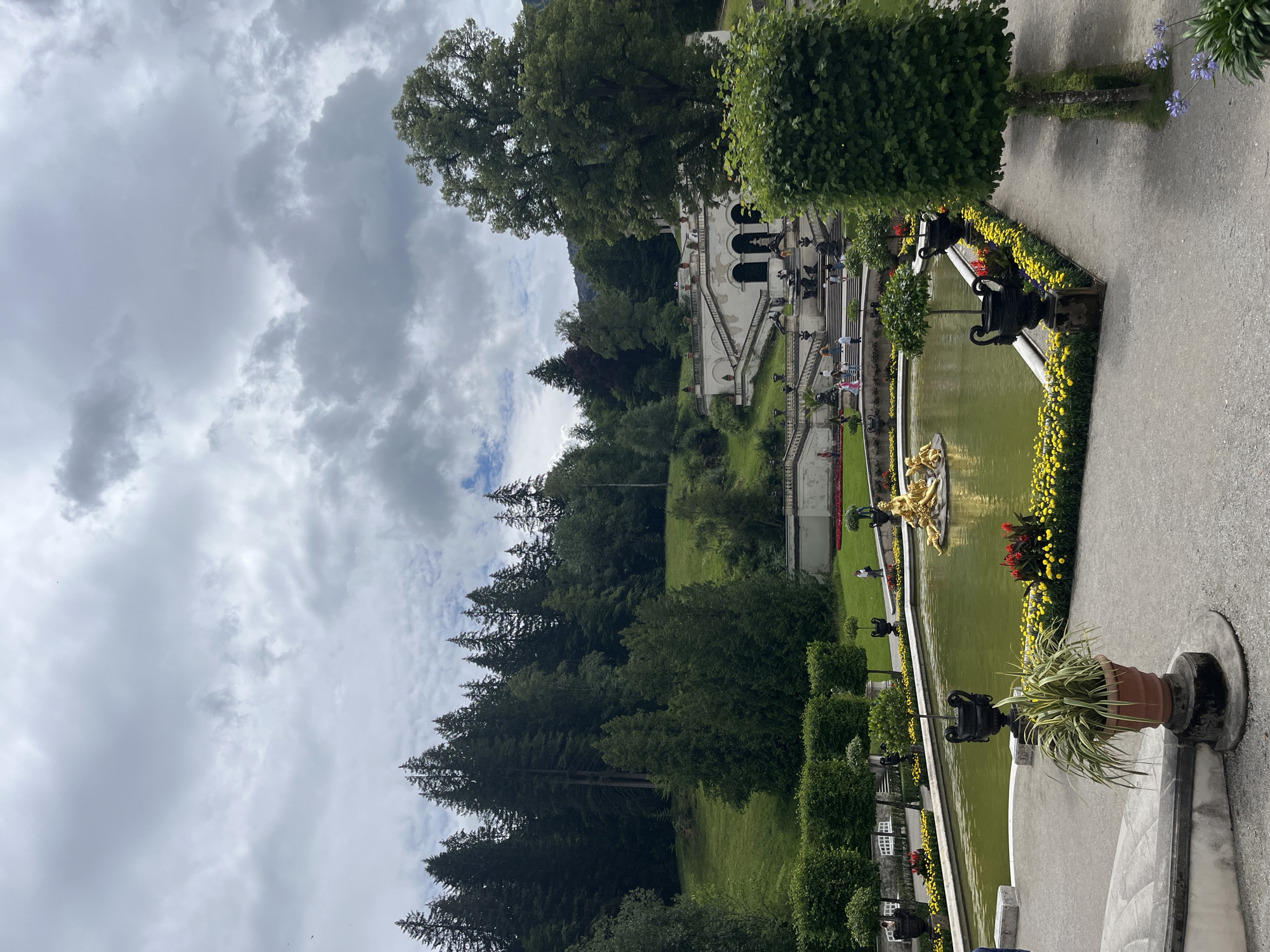 This image depicts the formal gardens of Linderhof Palace in Ettal, Germany. The scene is dominated by a large, rectangular pool with murky green water, featuring a golden sculpture in the center. The pool is bordered by a pathway and a vibrant display of yellow and red flowers. Several decorative black urns are strategically placed around the pool and along the pathway.
Behind the pool, an impressive tiered staircase with white balustrades leads up to an ornate white building with arched entrances. People are seen walking up and down the stairs and on the terraces. The background is filled with lush green lawns and a dense forest of tall evergreen trees, with a hill rising behind the palace structure. The sky is cloudy and overcast, suggesting a cool day.
In the foreground, a potted plant with long, spiky leaves sits on a stone pedestal, adding to the formal garden aesthetic. To the right, a tall, manicured tree stands beside a pathway. The overall impression is one of a grand, meticulously maintained European royal estate.