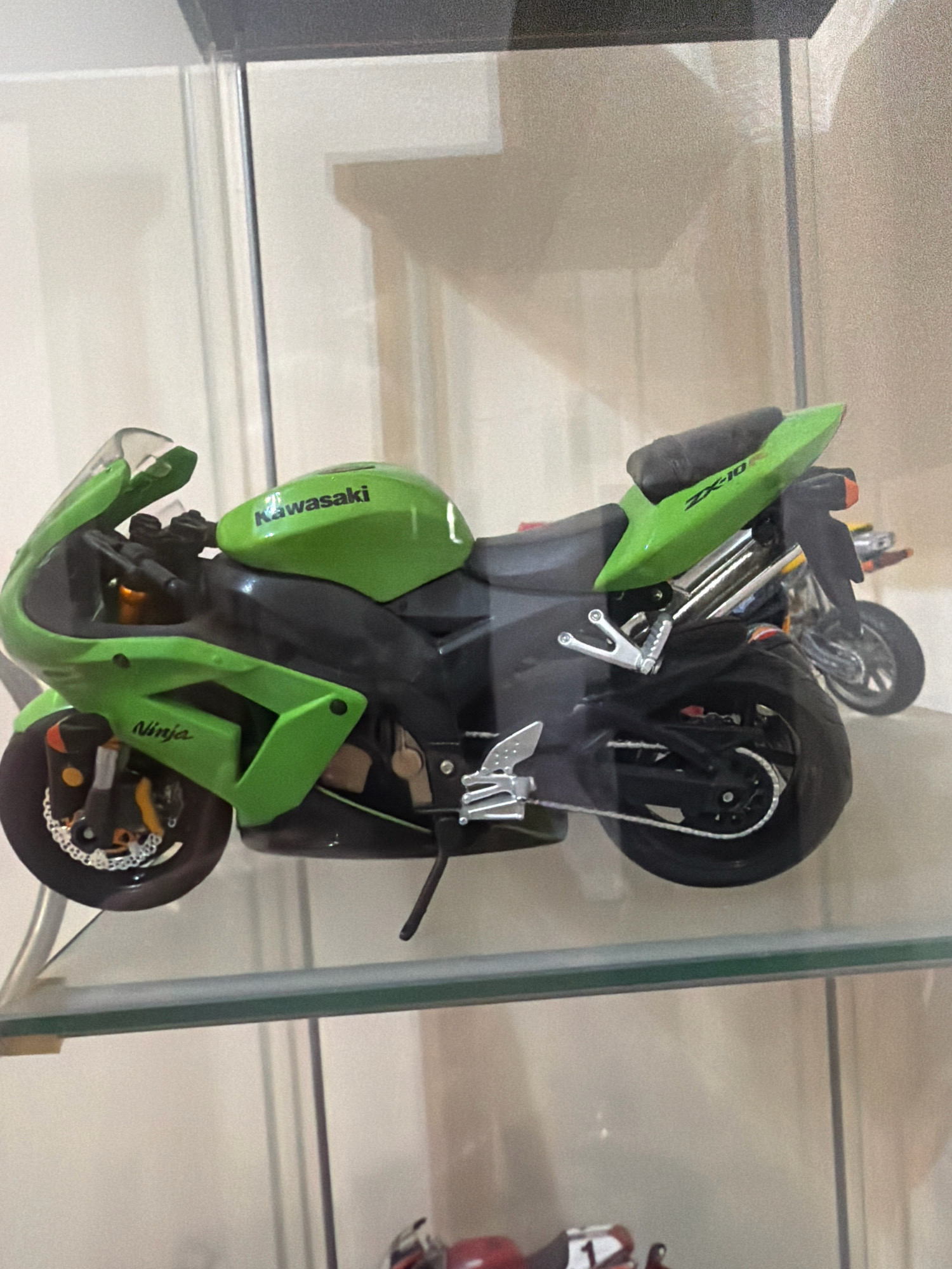 A close-up, medium shot captures a detailed green and black die-cast model of a Kawasaki Ninja ZX-10 motorcycle, displayed on a glass shelf within a display case. The motorcycle is positioned at a slight angle, facing left, with its front wheel closest to the viewer. The "Kawasaki" logo is visible on the fuel tank, and "Ninja" is printed on the side fairing. The rear of the motorcycle shows the "ZX-10" designation. Behind this model, another, less detailed motorcycle is partially visible. The background is out of focus, showing a neutral, light-colored wall with subtle shadows, suggesting an indoor setting. The lighting is even, highlighting the details of the motorcycle models. The scene is set within the Municipal Unit of Nafplio, Greece, specifically in a residential or private collection setting.