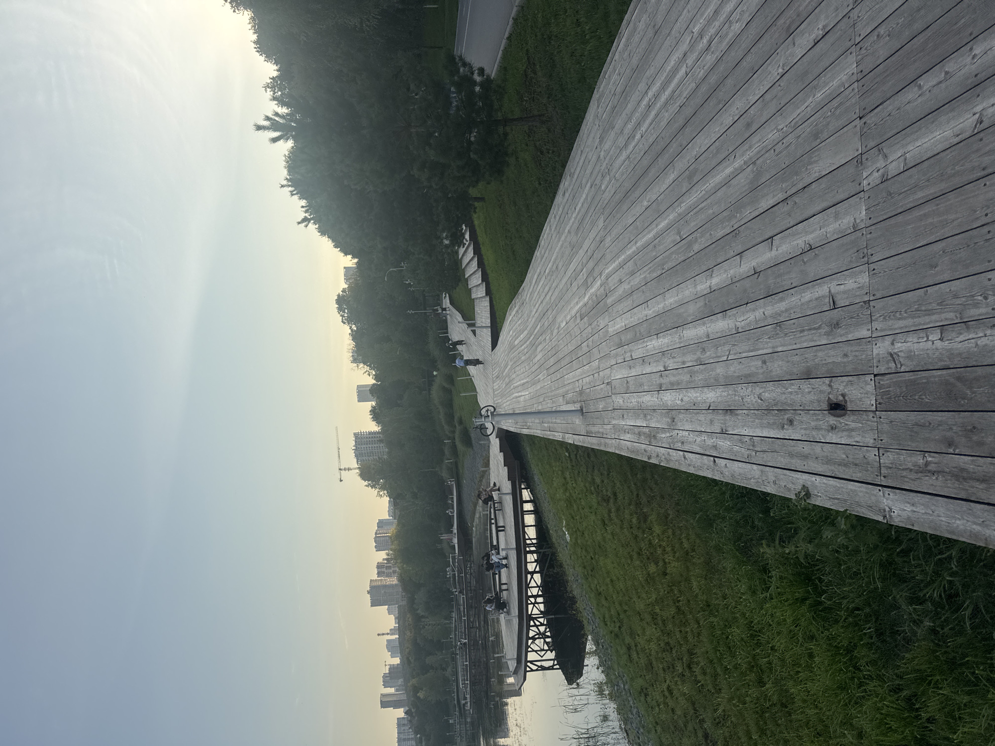The image depicts a wooden promenade extending into the foreground on the right side. To the left, a body of water is visible, with a wooden pier structure extending over it, featuring several individuals seated on benches. The pier is supported by visible struts. Beyond the water, lush green grass lines the bank, leading to a line of trees and a distant cityscape with numerous buildings and construction cranes. The sky is clear and pale blue. In the middle ground of the promenade, several individuals are seen cycling. A road is visible on the far right, bordered by trees and grass. The scene appears to be a park or recreational area within the city of Ufa, Russia, likely during daylight hours.