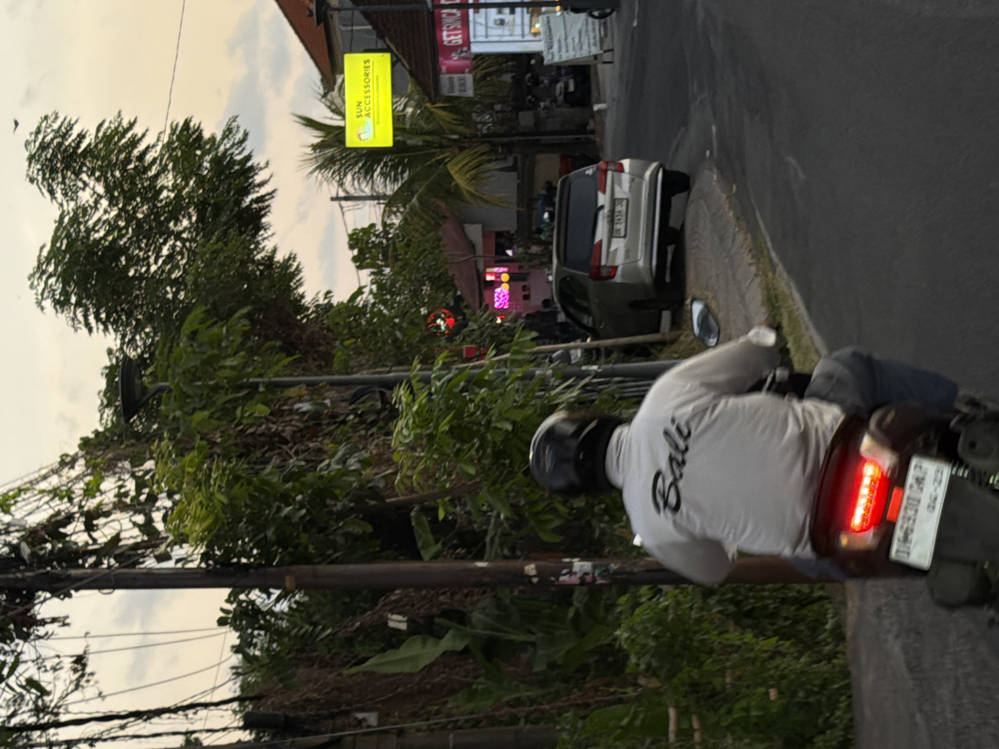 The image depicts an evening street scene in North Kuta, Indonesia. In the foreground, the back of a person wearing a black helmet, a white long-sleeved shirt with "Bali" printed in black script, and blue trousers is visible on a dark-colored motorbike. The motorbike's red taillight is illuminated, and its license plate "DK 6930 CAP" with "04-23" below it is legible. The motorbike is positioned on an asphalt road that curves slightly to the right, bordered by a concrete curb and a narrow strip of grass on the right.
To the left, a utility pole is heavily intertwined with wires and dense green tropical foliage. In the midground, further down the road on the right, a silver multi-purpose vehicle (MPV), identified by license plate "DK 1438 JD", is parked or moving. Beyond it, several buildings line the street. A bright yellow rectangular sign reading "SUN ACCESSORIES" with a small graphic is prominently displayed on the right. Further along the street, red and pink neon lights are visible from businesses on the left, and a partial sign starting with "GETSINC" is seen on a building on the far right. The background features an overcast sky with a warm, pale yellow-orange hue, characteristic of dusk or dawn. A single dark bird is visible in flight in the upper right portion of the sky. Lush green vegetation and distant structures complete the scene.