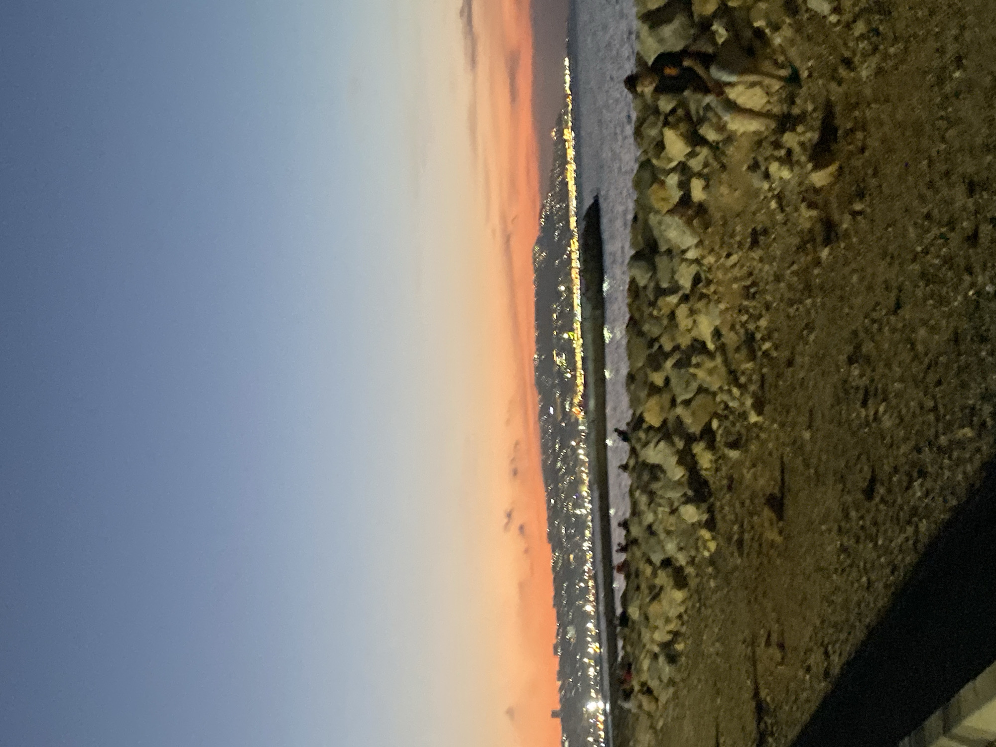 The image depicts a twilight scene of the coastline in Hussein Dey, Algeria. The sky transitions from a pale blue at the top to a vibrant orange and pink gradient near the horizon, indicating sunset or sunrise. In the distance, a city skyline is visible, illuminated by numerous lights, suggesting it is a developed urban area. The water is calm, with a slight ripple.
In the foreground, there is a rocky shoreline. A person, appearing to be a young male wearing a dark t-shirt and light-colored shorts, is seated on the rocks on the right side of the frame, facing away from the viewer. Other figures, possibly people gathered or walking, are faintly visible on the rocks closer to the water in the middle ground, some of whom appear to be sitting or standing. The ground in the immediate foreground is a mixture of dirt and gravel. The overall atmosphere is peaceful, with the natural light of the sky contrasting with the artificial lights of the city.