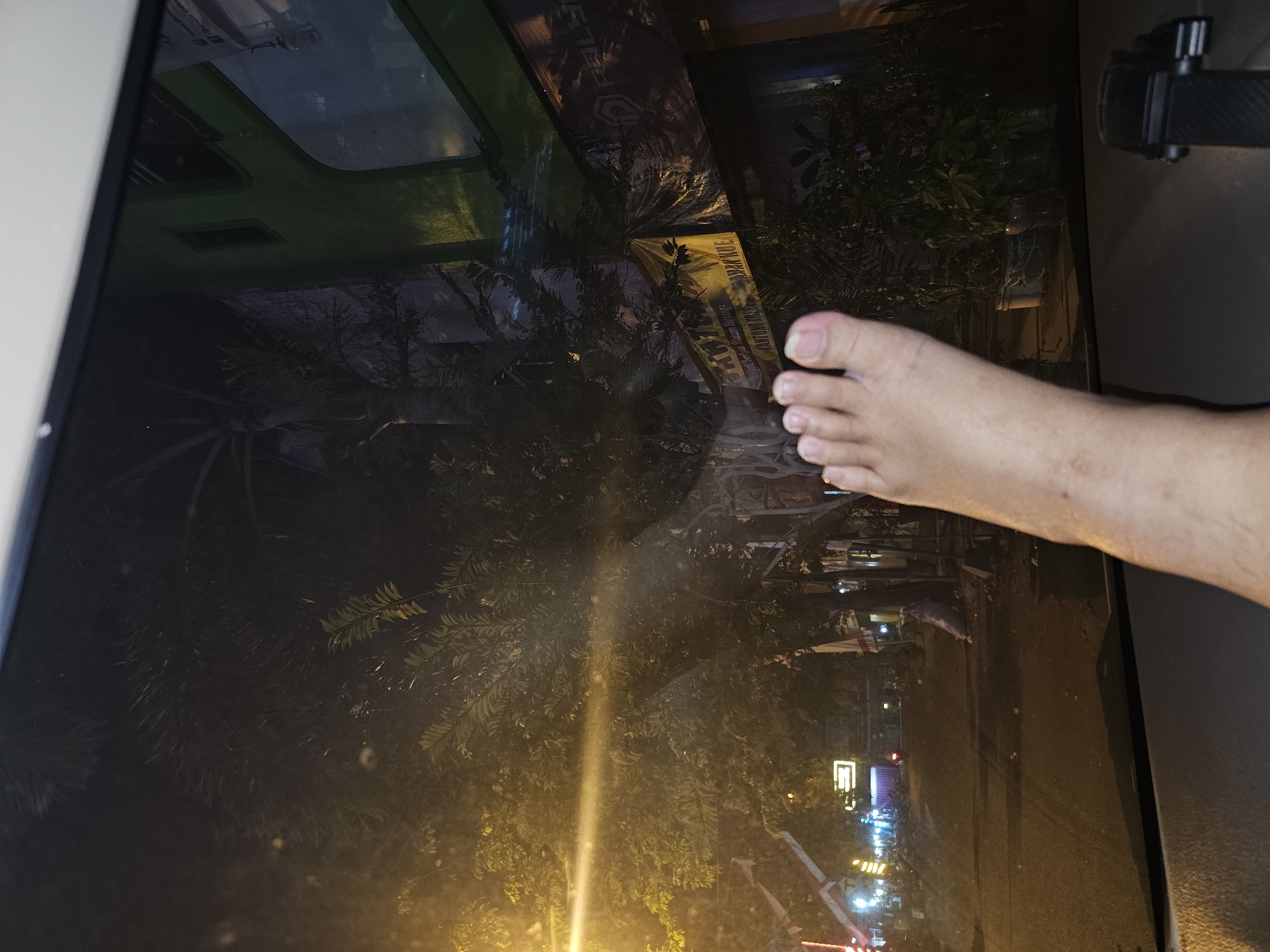 The image captures a nighttime street scene viewed through a car windshield, with a bare foot prominently positioned in the foreground on the dashboard. The foot, facing left, displays five toes. Beyond the windshield, a street is visible, illuminated by artificial lights and streetlights. Trees with dense foliage line the street, casting shadows. Several buildings are present, including a prominent green building on the right, reflecting the surrounding lights and foliage. A yellow banner with text, likely advertisements, is also visible on one of the buildings. The overall atmosphere suggests a warm, tropical night. The location is identified as the city of Lamongan, Indonesia.