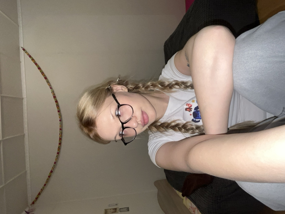 A woman with blonde hair in pigtails and glasses reclines on a couch. She wears a white t-shirt and grey pants. One arm is extended towards the camera, while the other rests on her leg. A colorful hula hoop leans against the wall in the background, adjacent to a white shelving unit.