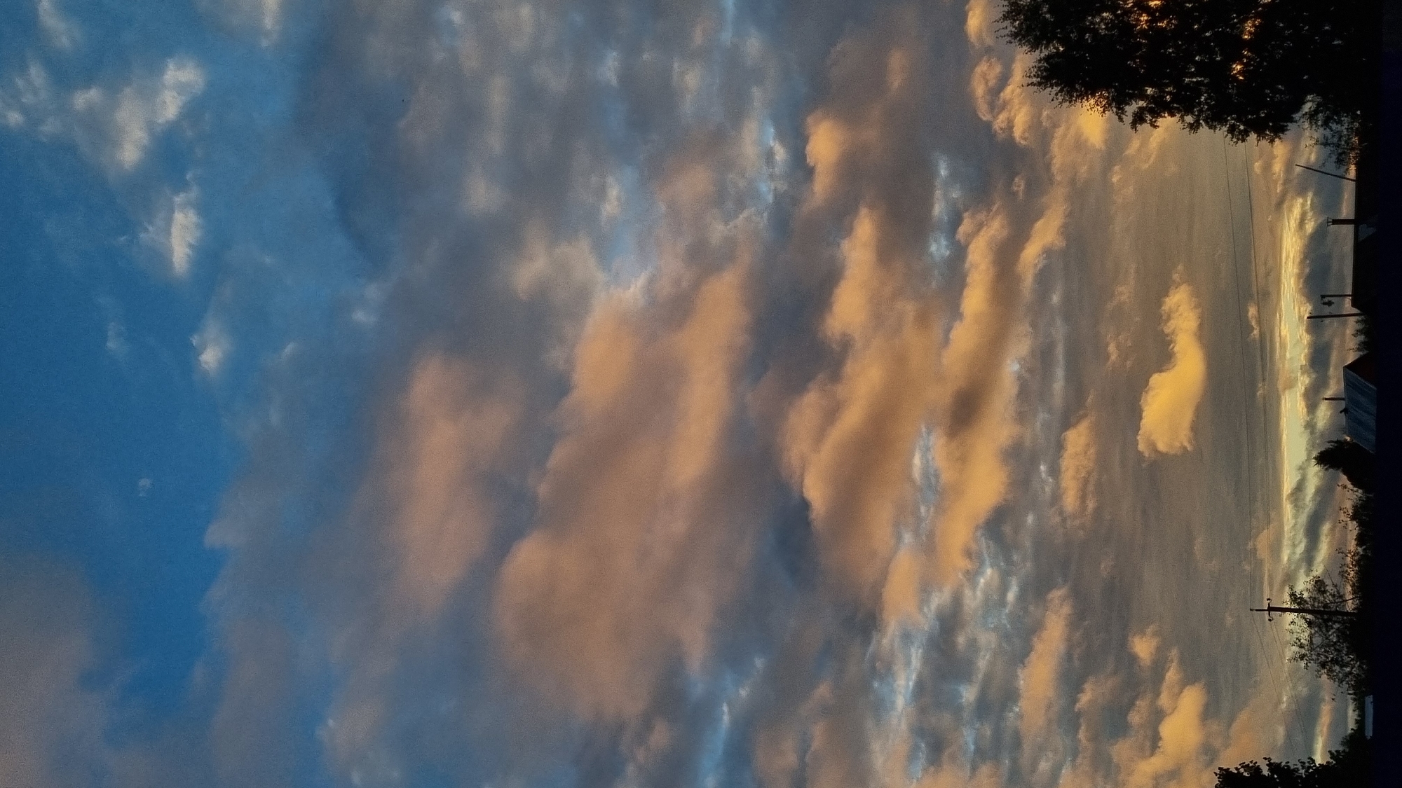 The image depicts a sky filled with scattered clouds tinted with orange and pink hues, likely from the setting sun. The sky transitions from a deep blue at the top to lighter shades near the horizon. In the lower portion of the image, a silhouetted line of trees and buildings forms the foreground. The architecture suggests a rural or suburban setting, with several rooftops visible. Electrical poles and wires are also present, indicating infrastructure. A single tree on the far right is prominently silhouetted against the sky. The scene is devoid of any people or discernable actions. The location is identified as Vysokoe, Russia.