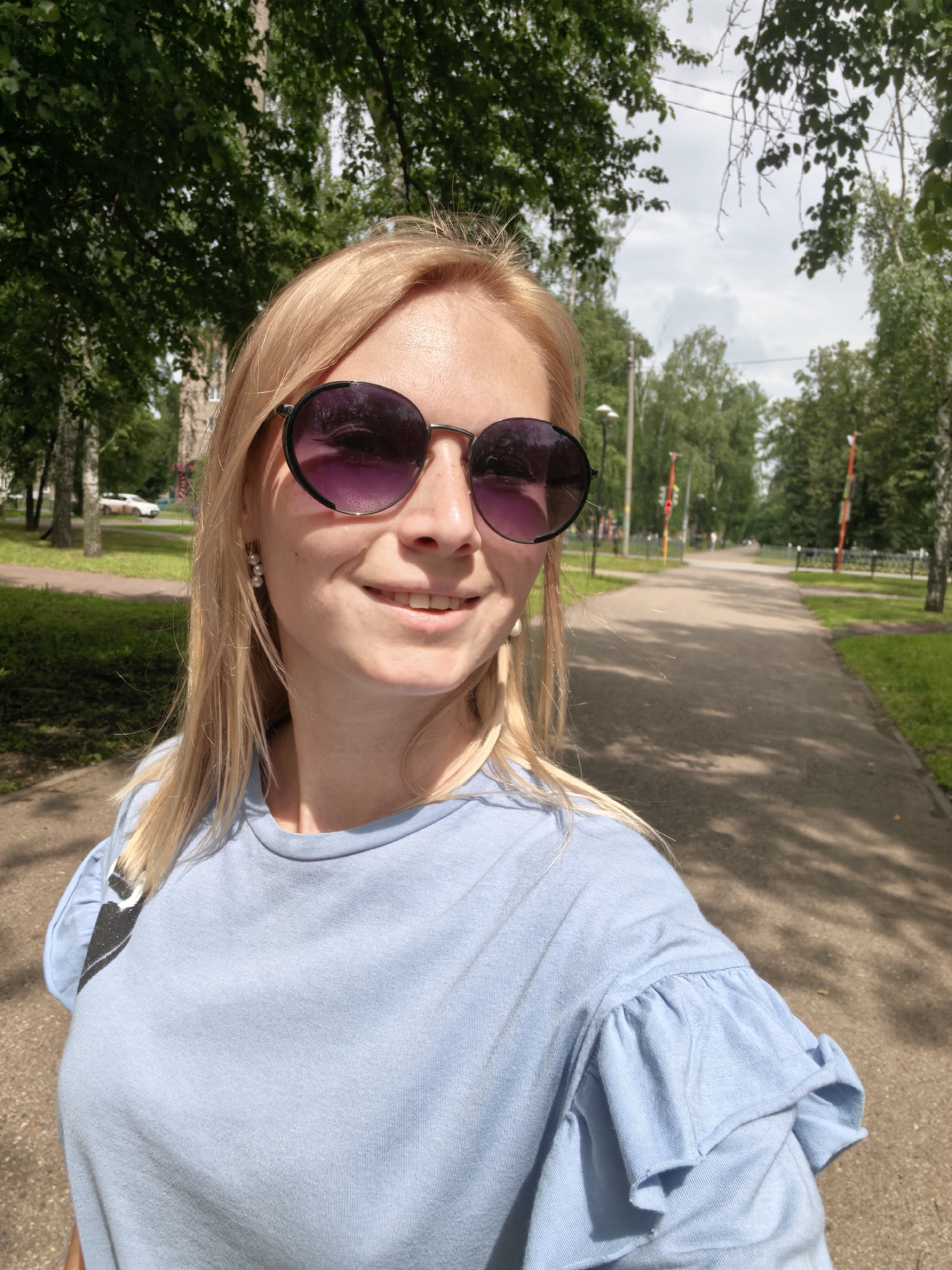 A blonde woman wearing a light blue t-shirt and large dark sunglasses is taking a selfie outdoors in a park on a sunny day. She has a slight smile on her face and is wearing pearl earrings. Behind her, a paved path winds through a tree-lined park. Several tall, colorful poles are visible along the path in the distance. The overall environment suggests a pleasant day for a walk in a public green space.