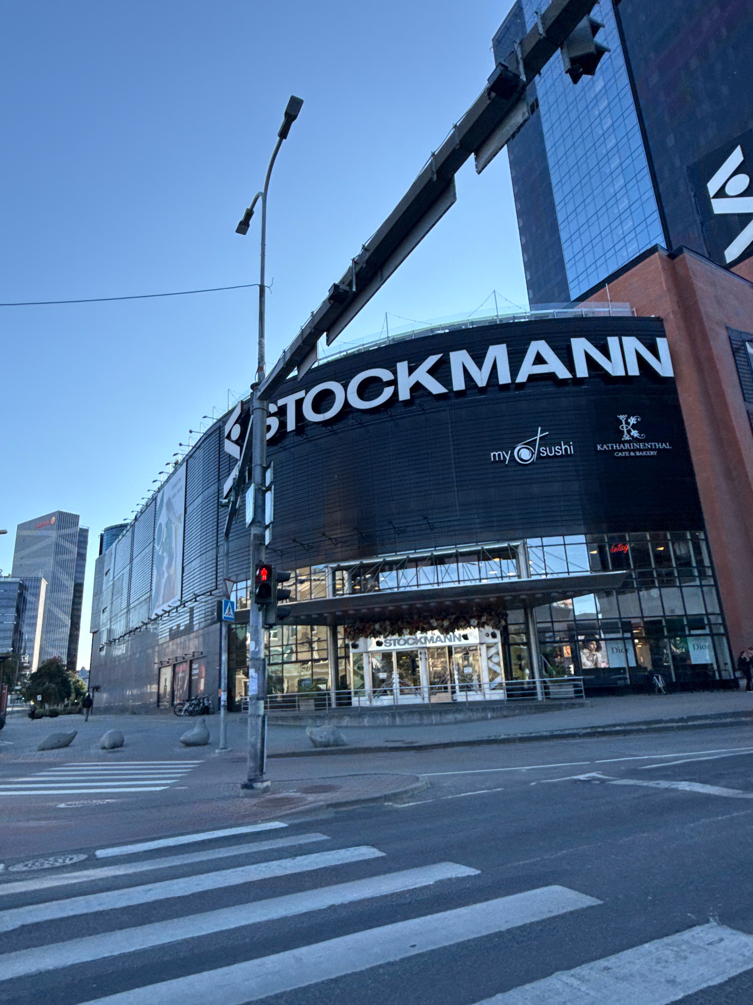 The image shows the Stockmann department store in Tallinn, Estonia, located on the corner of Liivalaia and Gonsiori streets. The building's facade is predominantly black, featuring a large "STOCKMANN" logo in white lettering. Above the entrance, the "my sushi" and "KATHARINENTHAL CAFE & BAKERY" logos are visible. The street in front of the store has a crosswalk with white stripes. A traffic light is visible, showing a red signal. Modern glass-fronted buildings, including the adjacent Solaris Keskus, are in the background. A street lamp with multiple fixtures stands in the foreground. The sky is clear and blue. There are a few pedestrians and parked bicycles visible in the background.