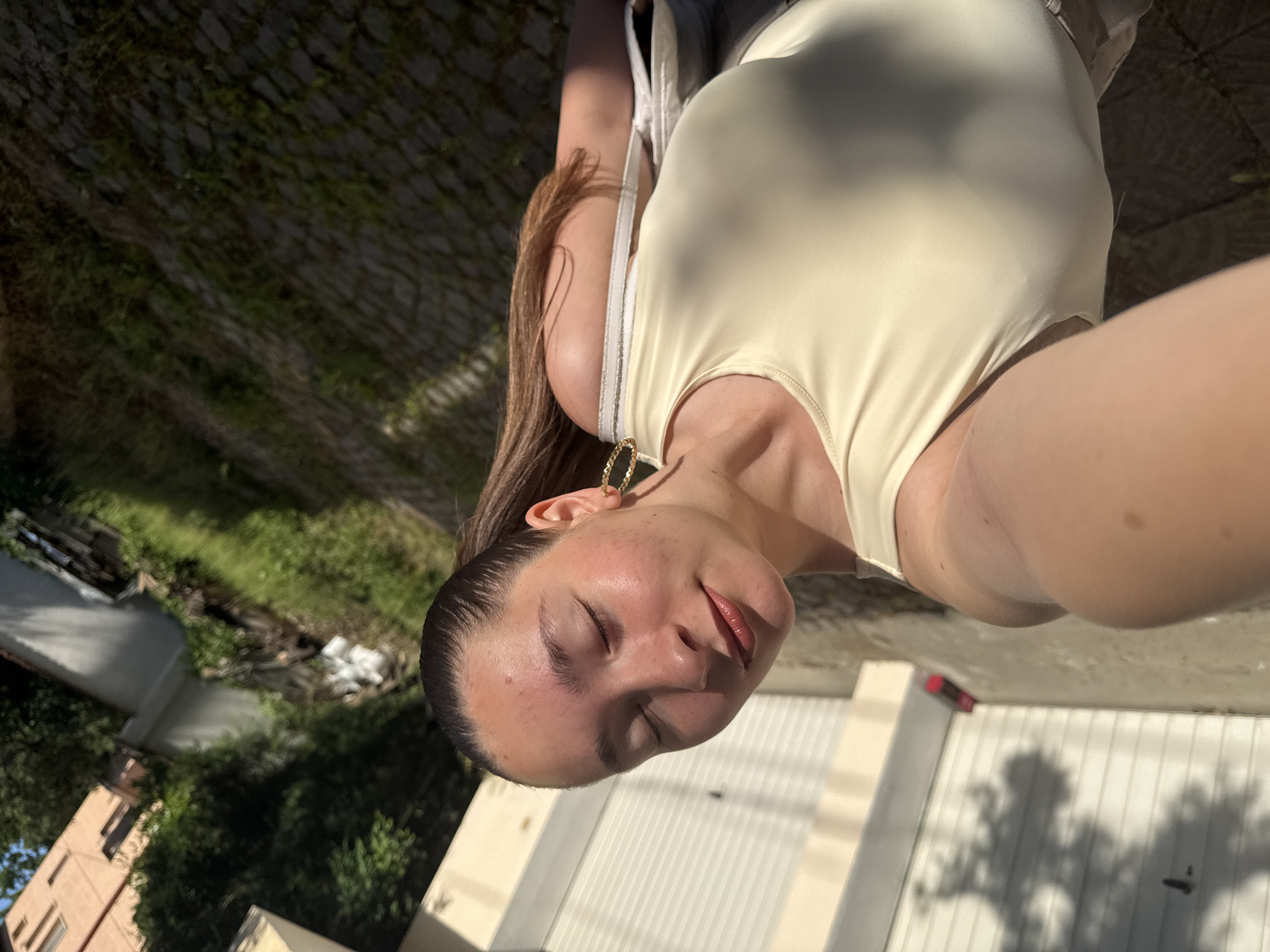A young woman with her eyes closed and head tilted back is taking a selfie outdoors. She is wearing a cream-colored tank top and large gold hoop earrings. Her hair is pulled back into a ponytail. The background shows a paved area with grass growing between the stones, a building with garages, and trees. The image appears to have been taken in the city of Sofia, Bulgaria. The lighting suggests it is either early morning or late afternoon.