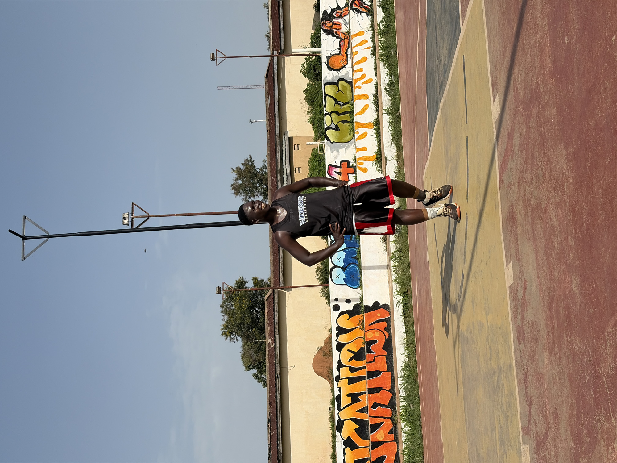 The image captures a man standing on a basketball court in Jos, Nigeria. The court's surface is divided into colored sections: red, yellow, and dark. He is wearing a dark sleeveless top with "WIDENER BASKETBALL" printed on it, along with black shorts with red trim and athletic shoes. His hands are on his hips. A light-colored wall with graffiti runs along the edge of the court, featuring various colorful designs. Behind the wall is a building and greenery, including trees. A basketball hoop structure is visible against a clear, blue sky.