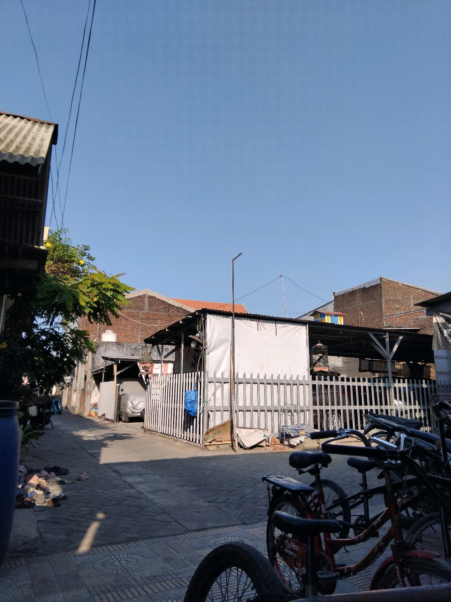 The image depicts a street scene in Kepuh Kiriman, Indonesia, under a clear blue sky. In the foreground, several bicycles are parked, some with red frames. The street itself is paved with a patterned surface. To the left, a large blue cylindrical object is partially visible, along with scattered footwear. Further down the street, a white fence encloses several structures, including a building with a white tarp-covered roof and a car covered in a grey tarp. Brick buildings with orange tiled roofs are visible behind this structure. The environment appears to be a residential or mixed-use area, characterized by modest buildings and everyday objects. The lighting suggests it is daytime, likely morning or afternoon, with shadows cast across the pavement. No people are clearly visible in the scene.