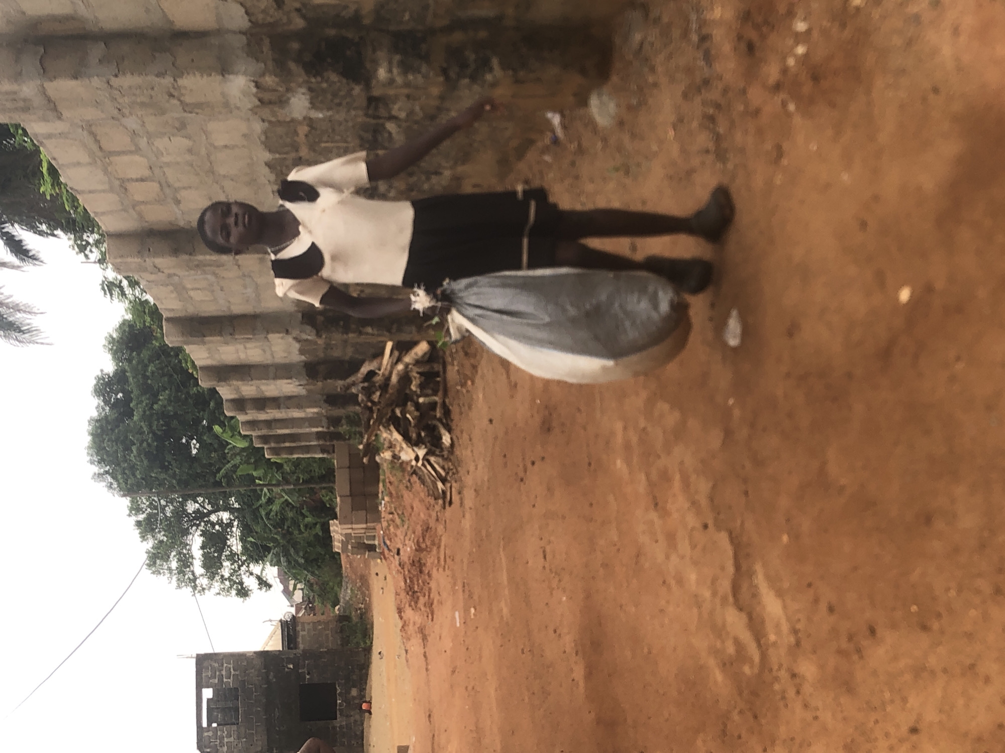 This image from Ukwu-Abwa, Nigeria, shows a young person standing outdoors on a reddish-brown dirt path. The individual, who appears to be a child or teenager with dark skin, is wearing a white short-sleeved top with black accents and black shorts. They are holding a large, grey and white bag, which appears full and bulky, in front of them. The person is looking directly at the camera with a neutral expression.
The background features a structure made of unrendered concrete blocks, possibly an unfinished building or wall. To the left, lush green trees and foliage, including what looks like palm fronds, are visible against an overcast sky, suggesting a cloudy day. A pile of wooden debris or branches lies on the ground behind the person. Thin wires, potentially power or communication lines, traverse the upper left side of the frame. The overall scene depicts a rural or semi-urban environment with ongoing construction or development. There is no visible text in the image.